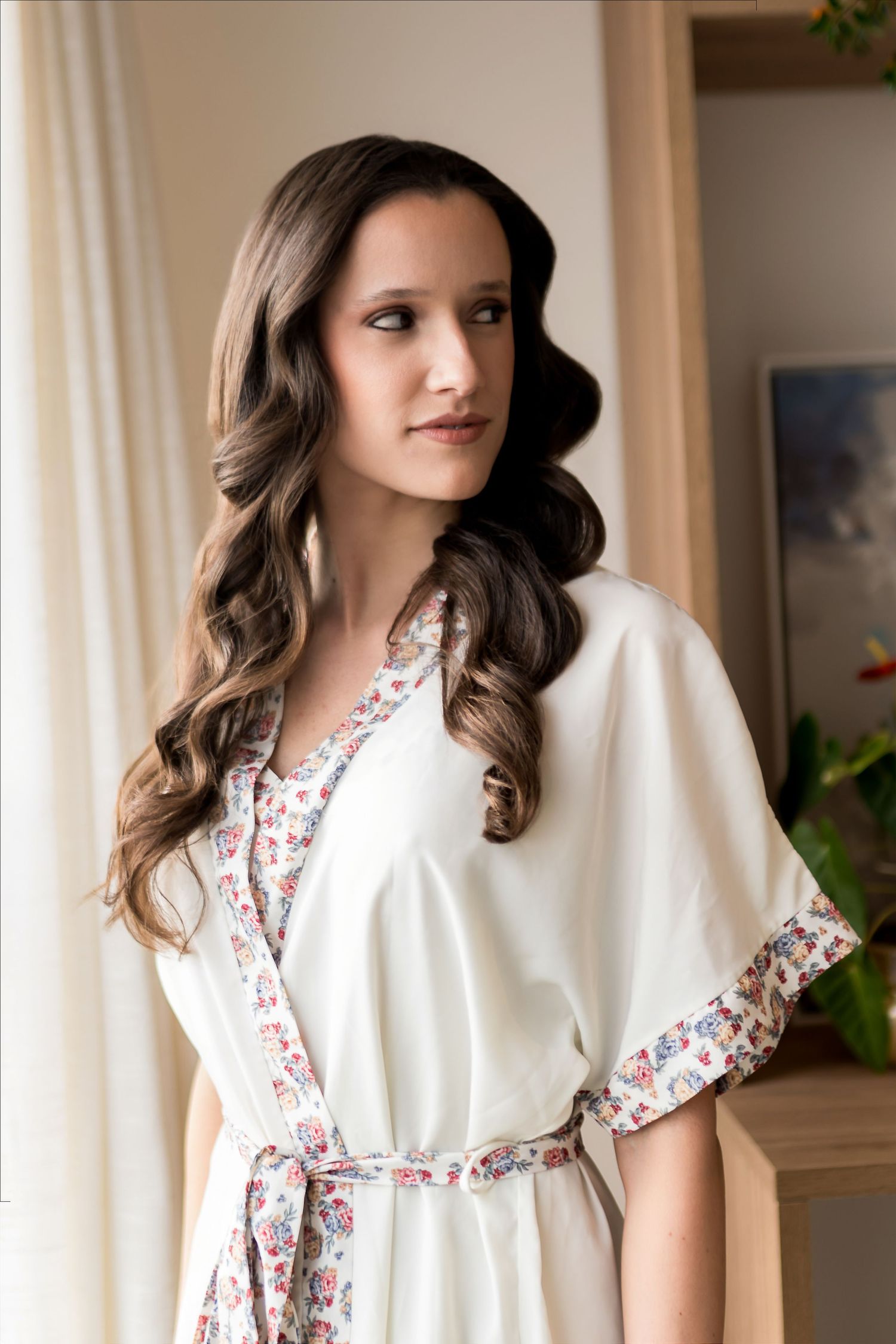 Woman in white floral-trimmed robe with long curly brunette hair, elegant bridal getting-ready look.