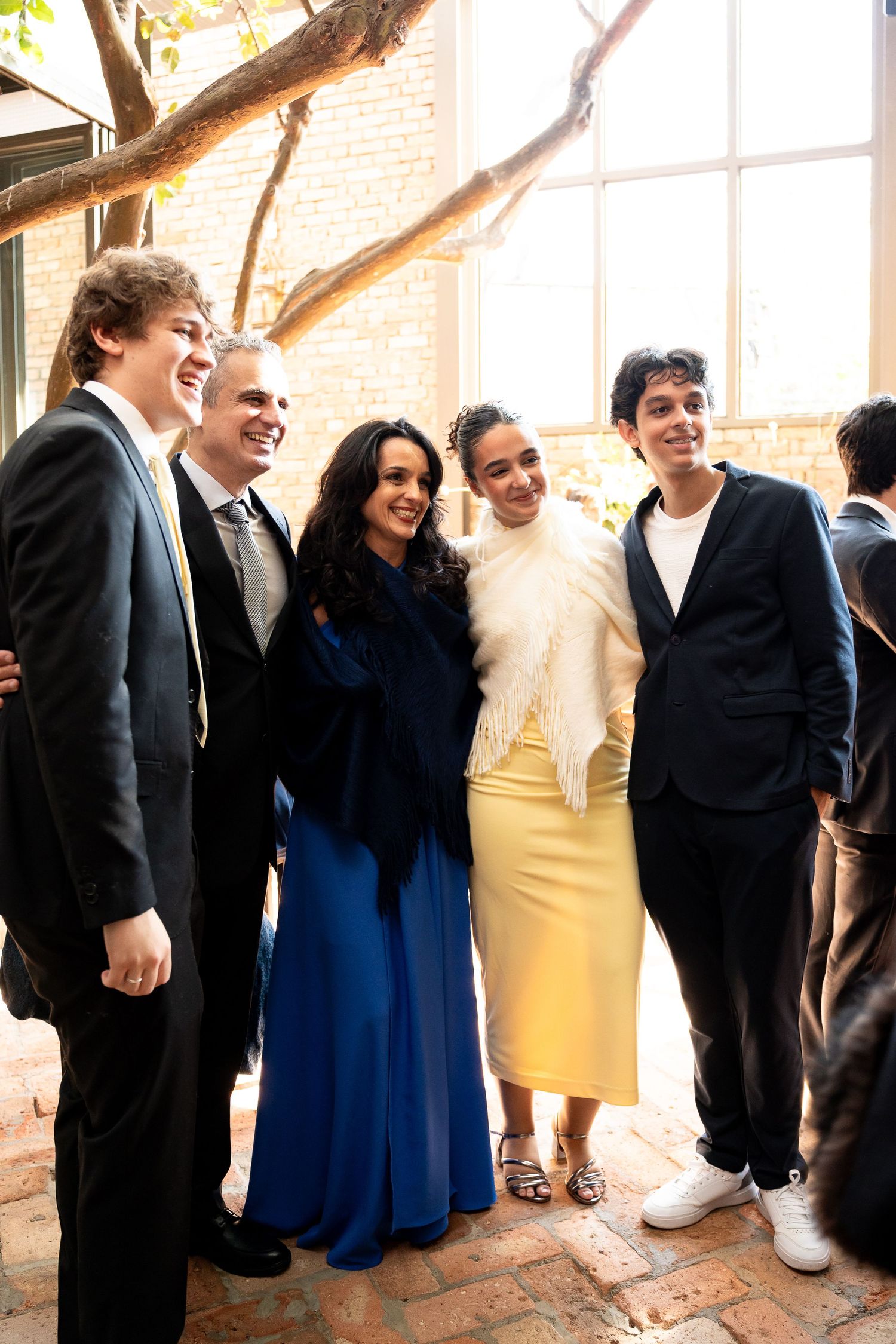 Group of five elegantly dressed people smiling together indoors near a large window and brick wall.