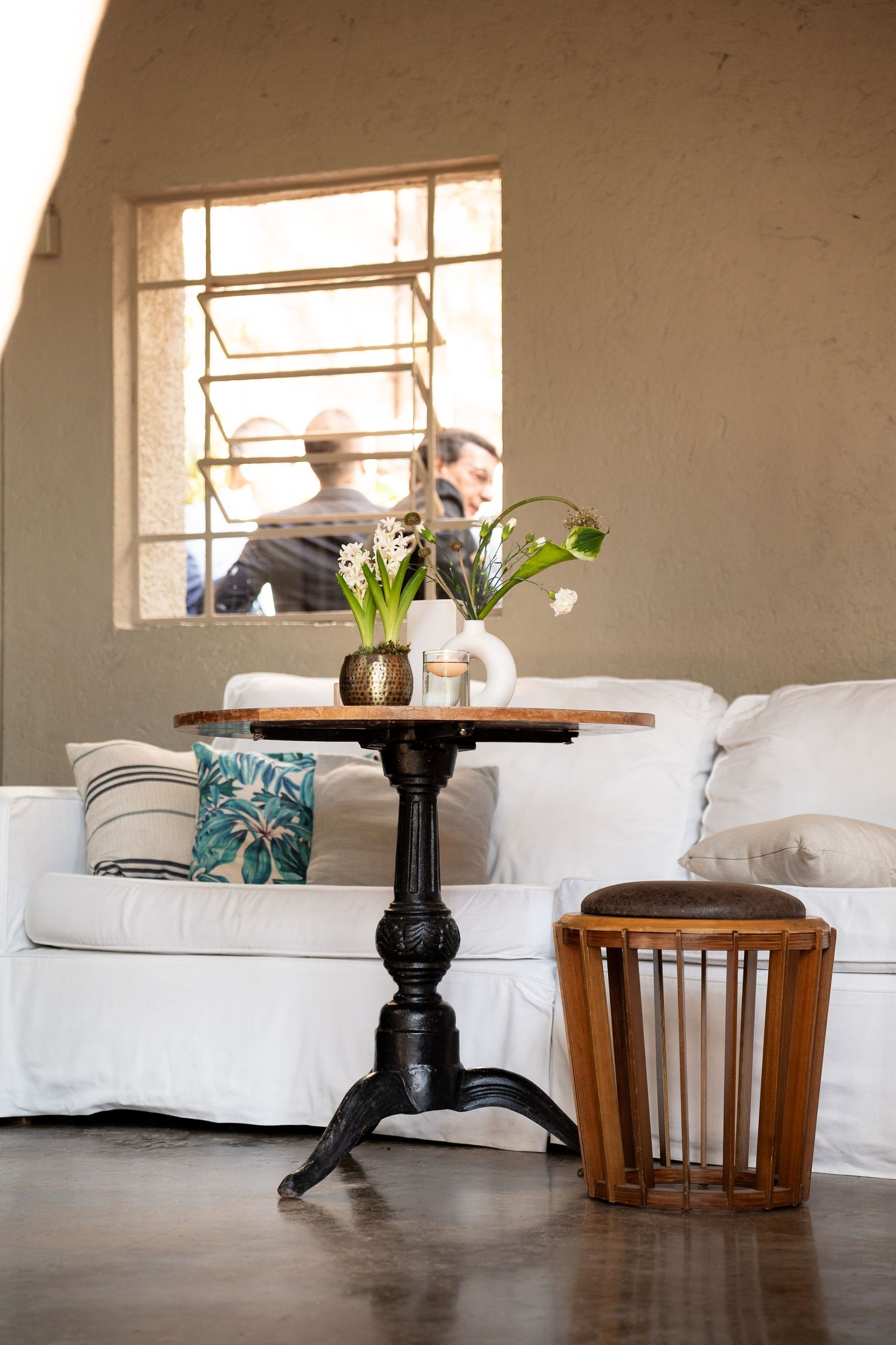 Cozy interior with white sofa, black pedestal table, fresh flowers, and wooden stool near a sunlit window.