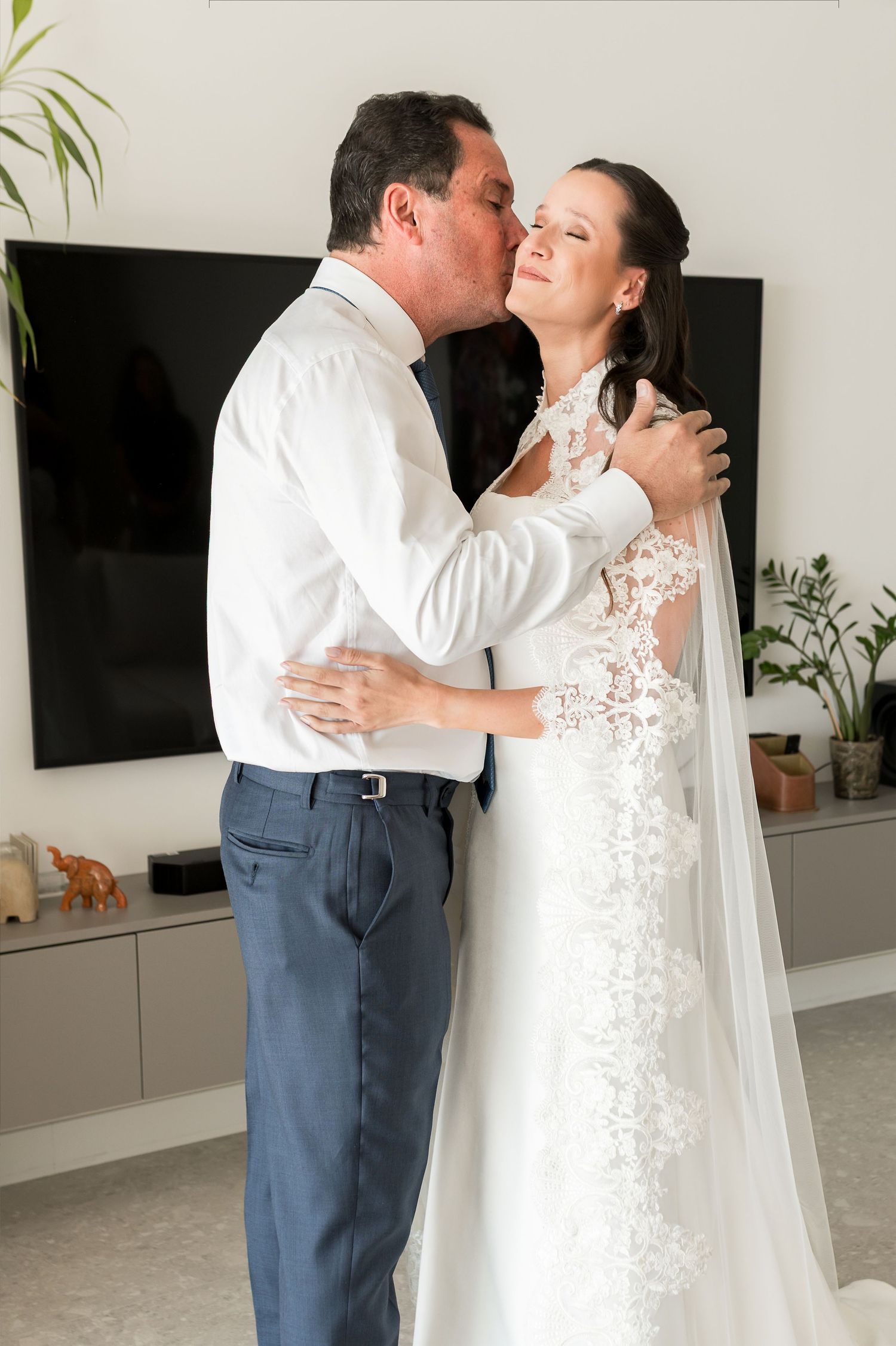 A groom kisses a smiling bride in a lace wedding dress and veil in a modern living room.