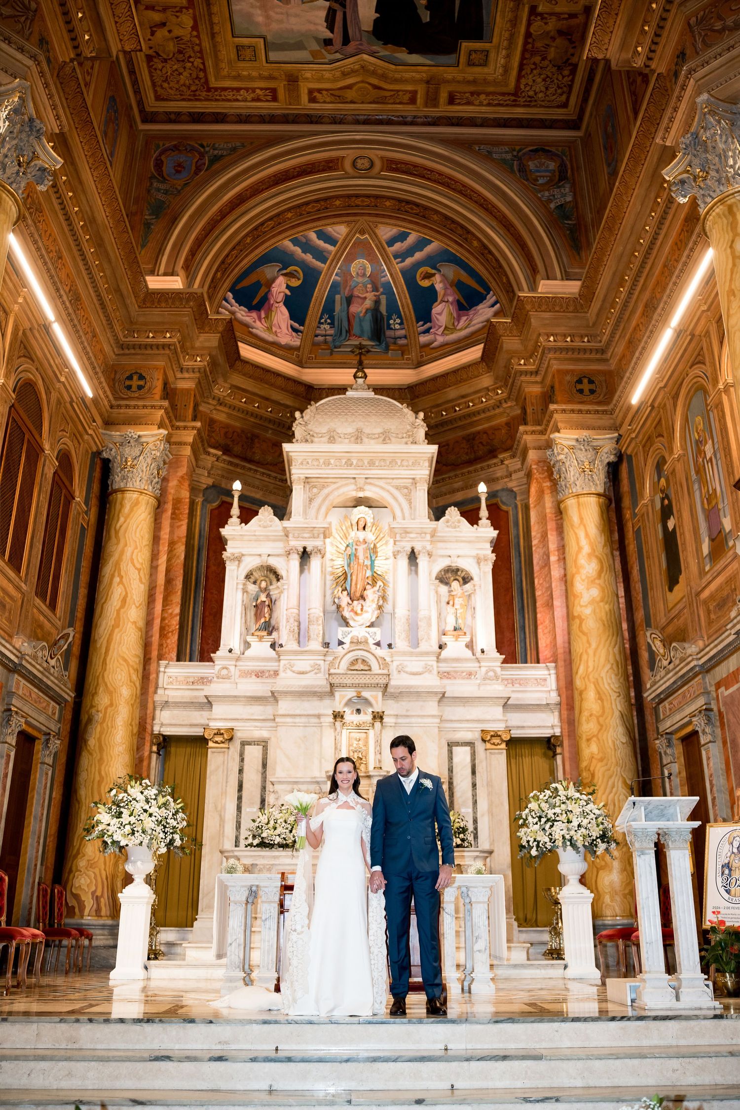 Bride and groom stand at ornate church altar with marble decor, floral arrangements, and painted ceiling during wedding ceremony.