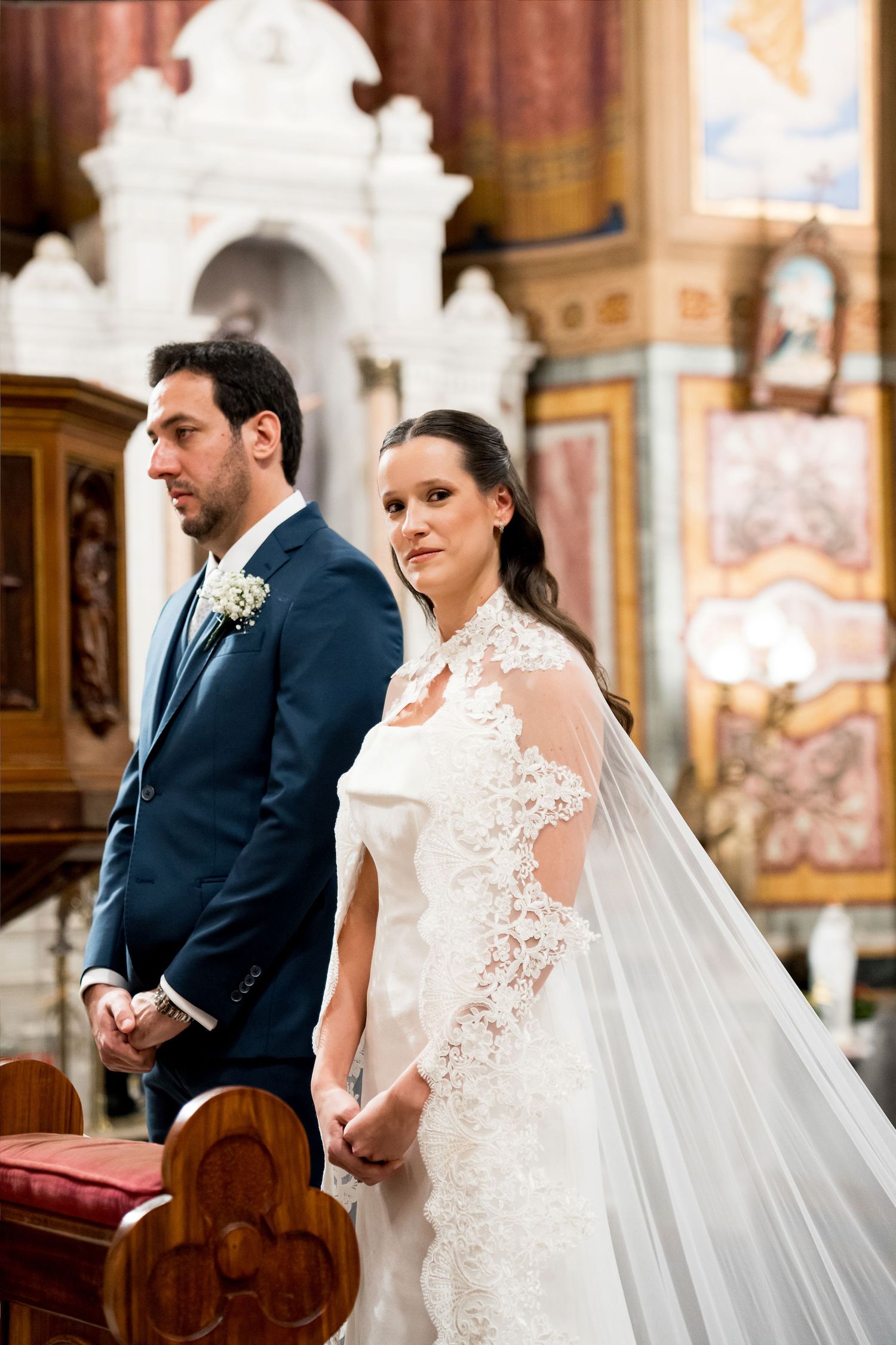 Bride in lace gown with veil and groom in navy suit standing at church altar during wedding ceremony.