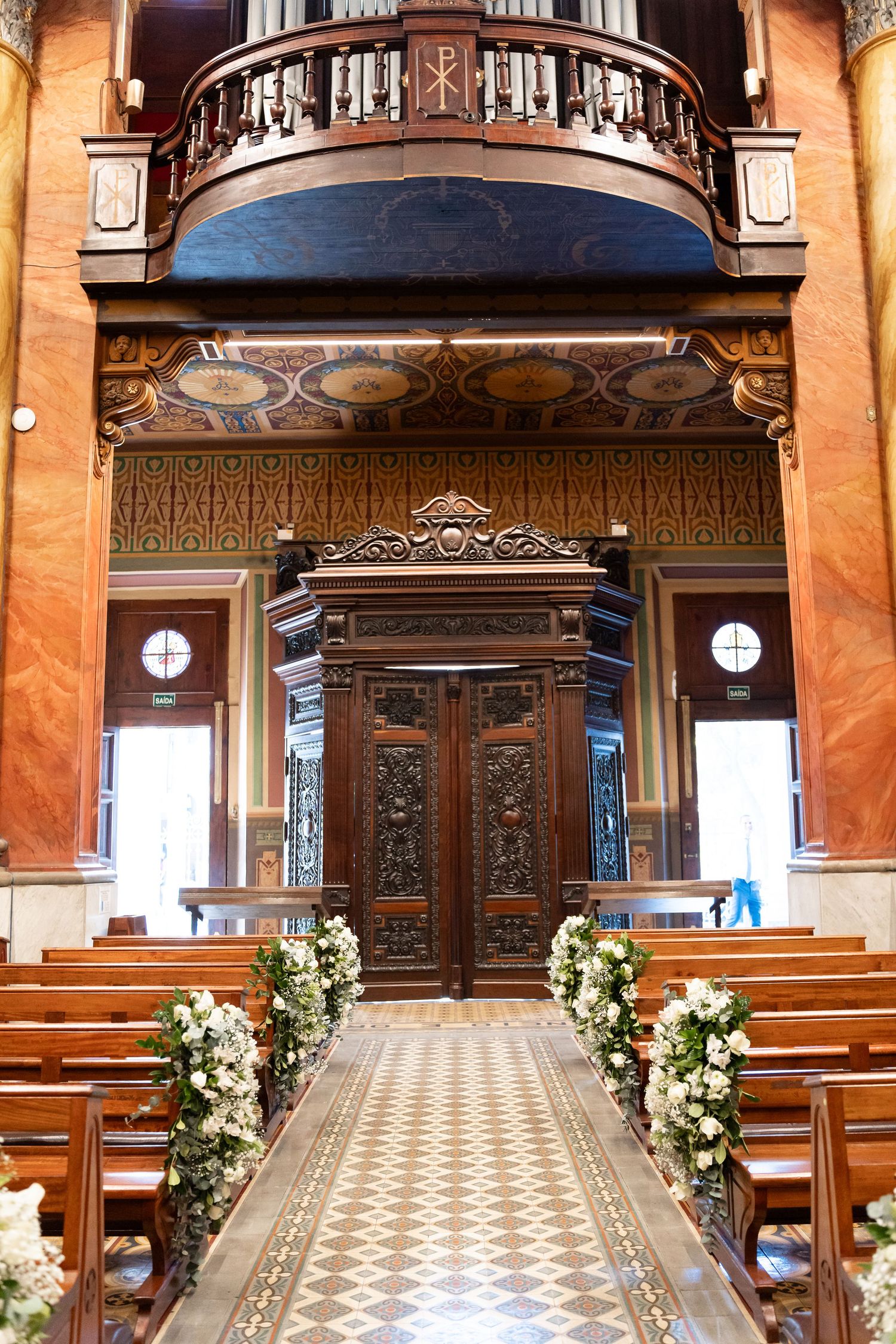 Elegant church interior with wooden pews adorned with white floral arrangements leading to ornate doors and a choir loft.