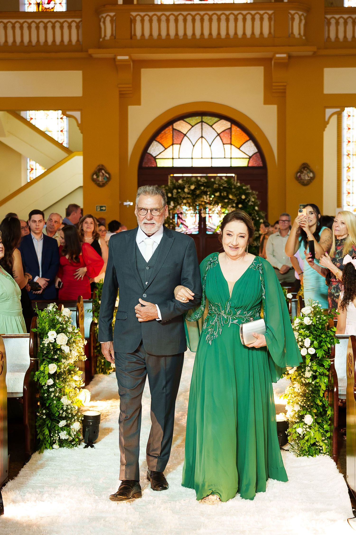 Elegant couple walks down church aisle, woman in green gown, man in suit, guests watching at wedding ceremony.