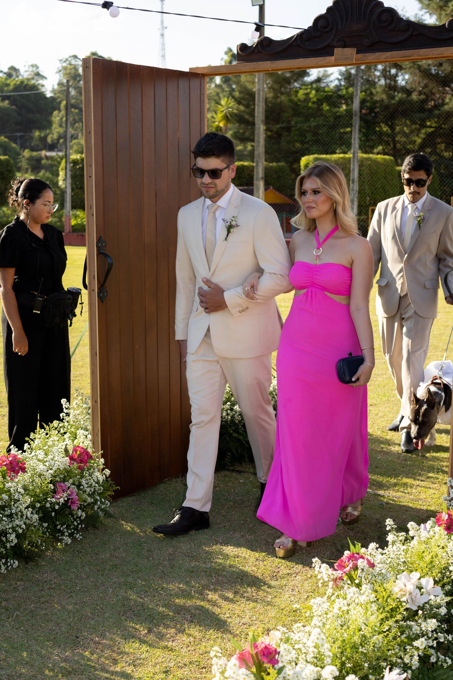 Couple in formal attire walking through floral arch at outdoor wedding, man in cream suit, woman in pink gown.