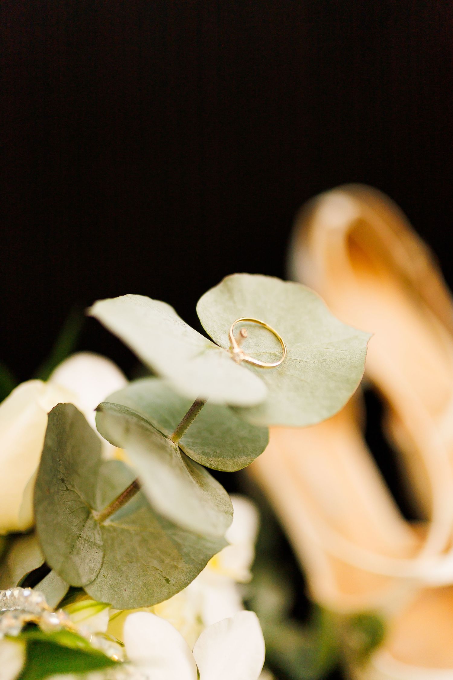 Gold wedding ring resting on eucalyptus leaves with white flowers and blurred bridal heels in the background.