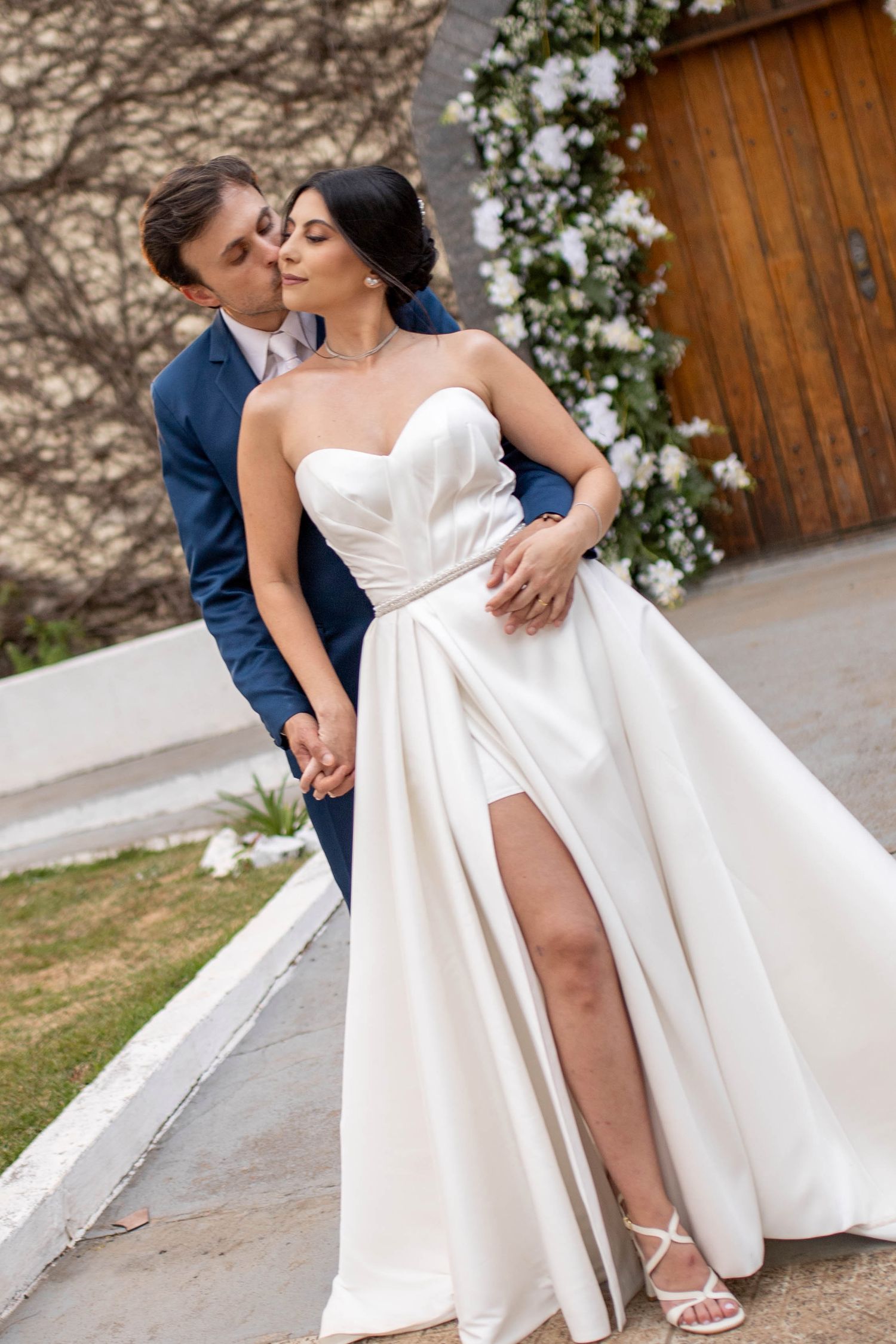 A couple in wedding attire shares a romantic moment, groom in blue suit kissing bride in white strapless gown outdoors.