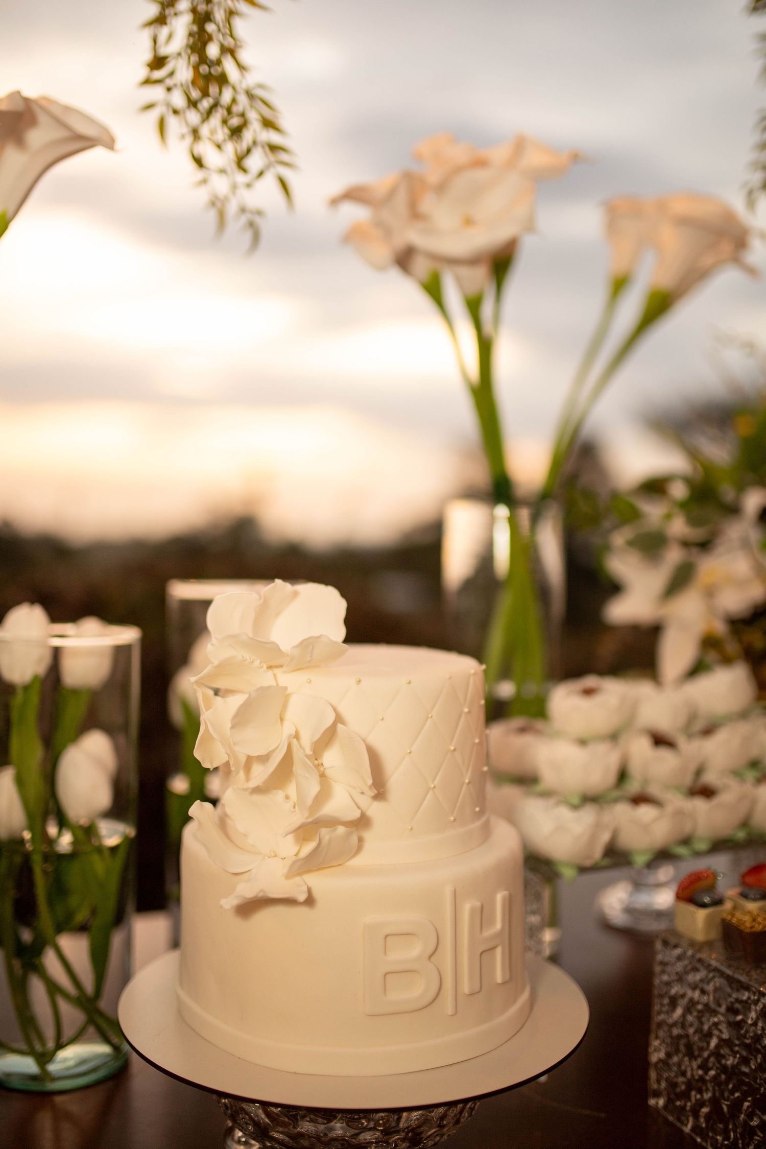 Elegant two-tier white wedding cake with monogram letters and calla lily flowers on a decorated dessert table at sunset.