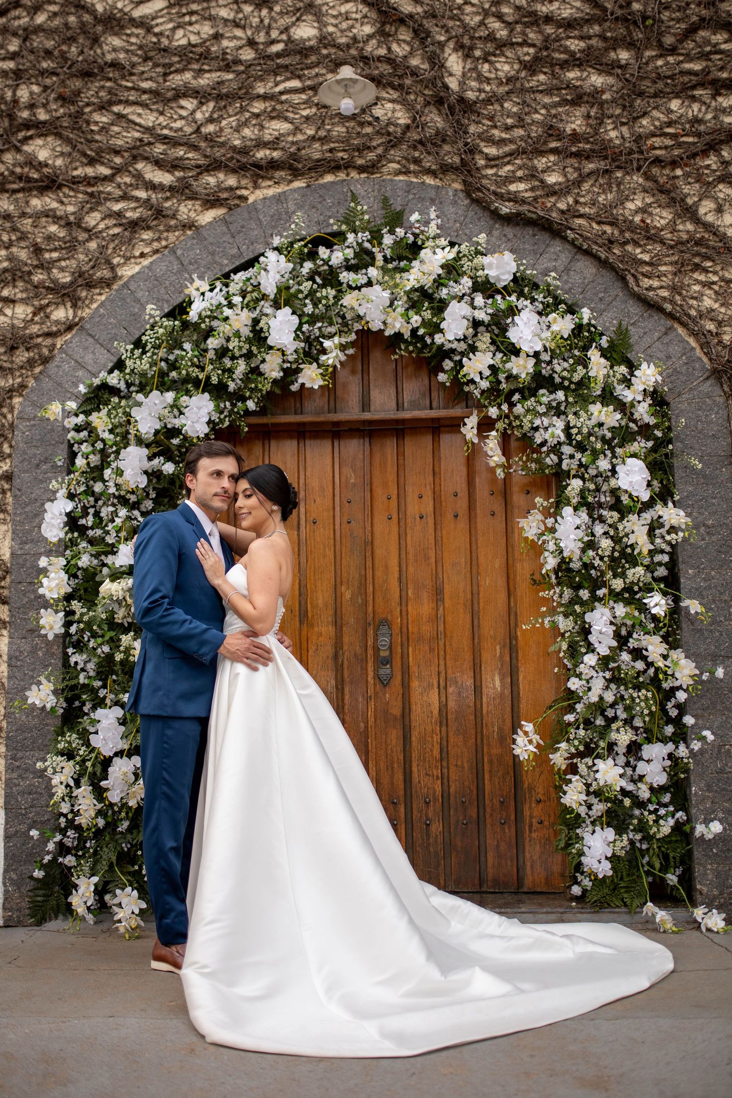 Bride and groom embracing under a floral arch with white flowers at a wooden door wedding venue.
