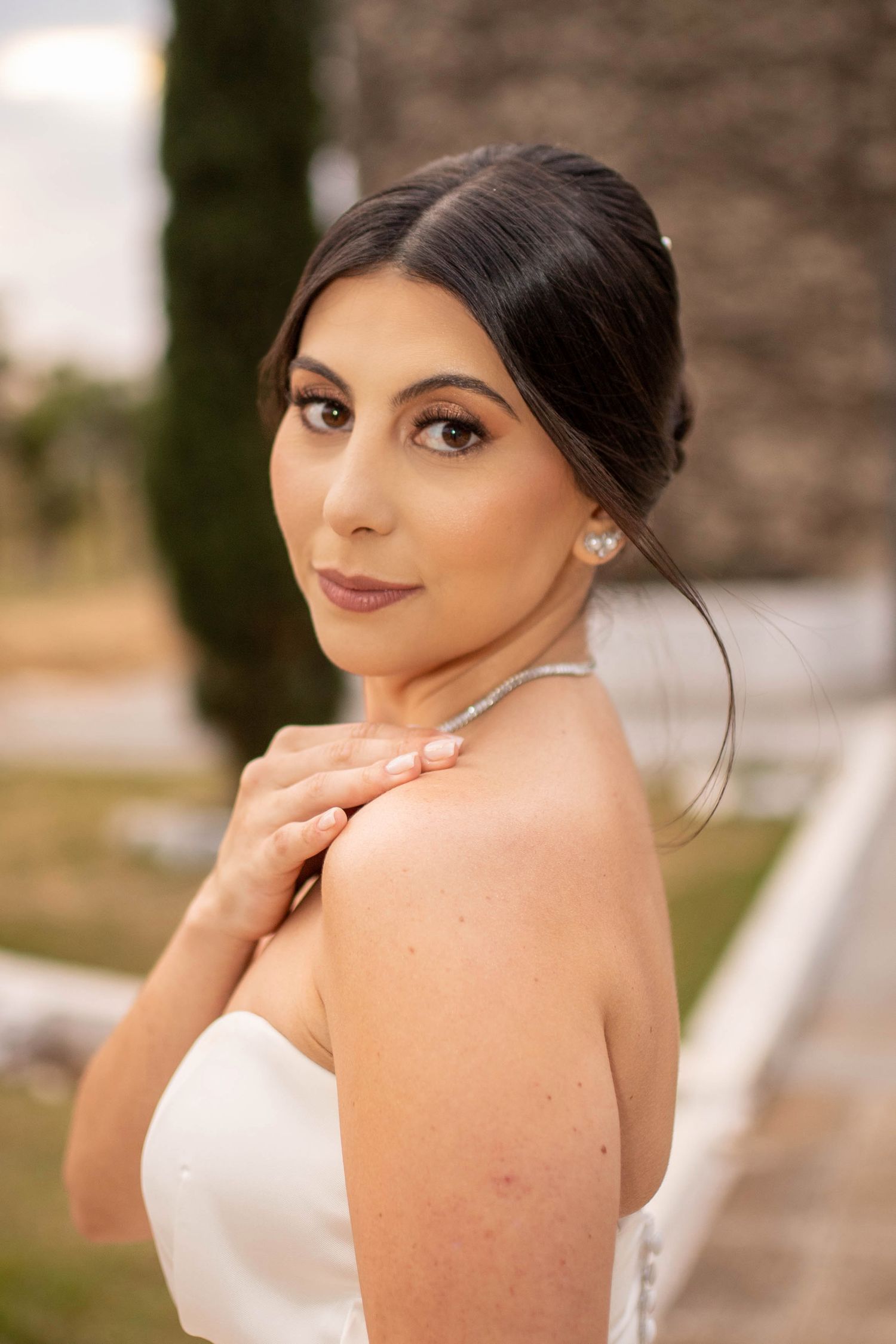 A bride in a white strapless gown poses gracefully, wearing pearl earrings and a delicate necklace outdoors.
