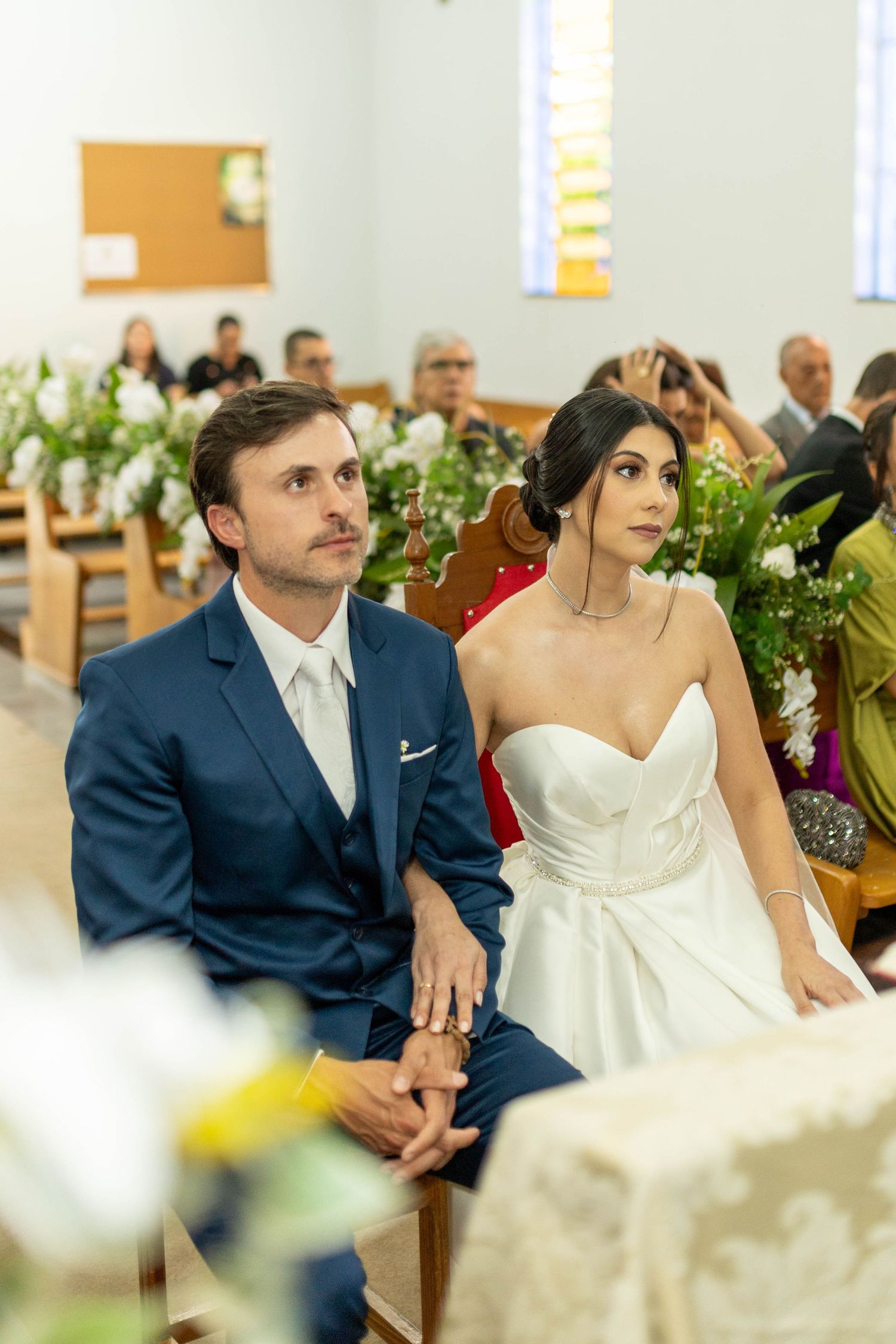 Bride and groom sitting together at church altar during wedding ceremony, bride in strapless white gown, groom in navy suit.
