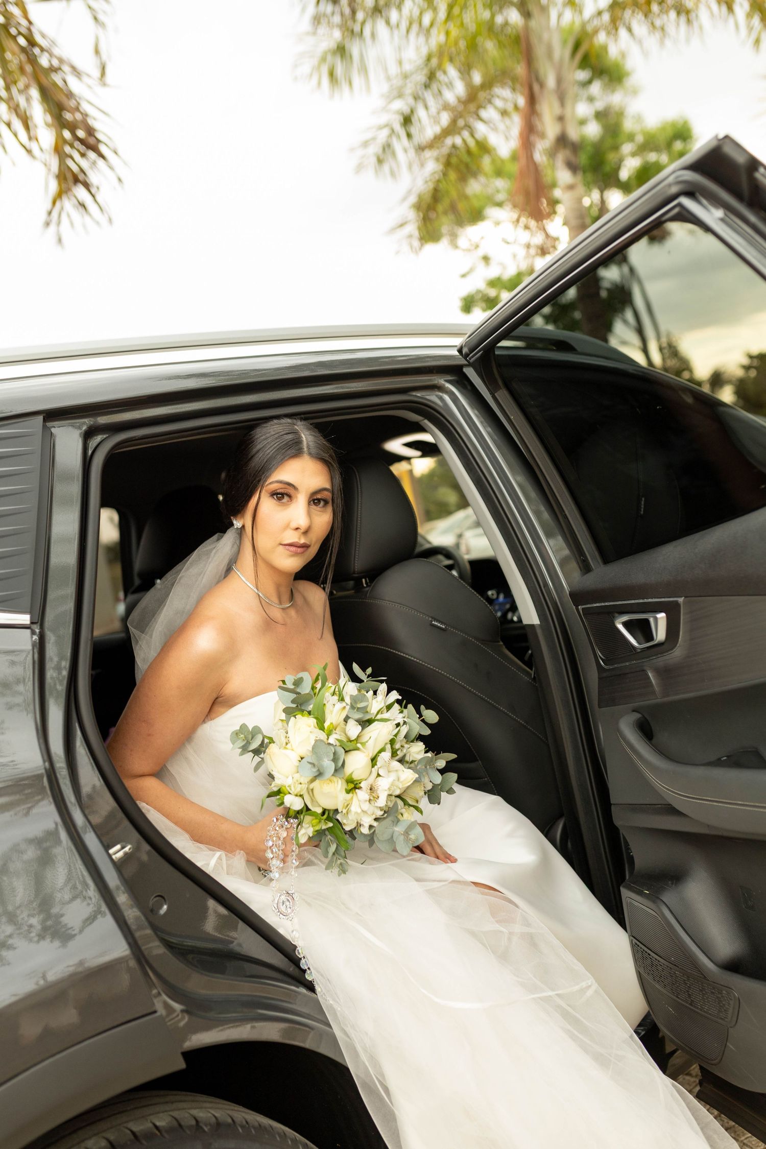Bride in white strapless gown holding bouquet of white flowers while seated in a car on her wedding day.