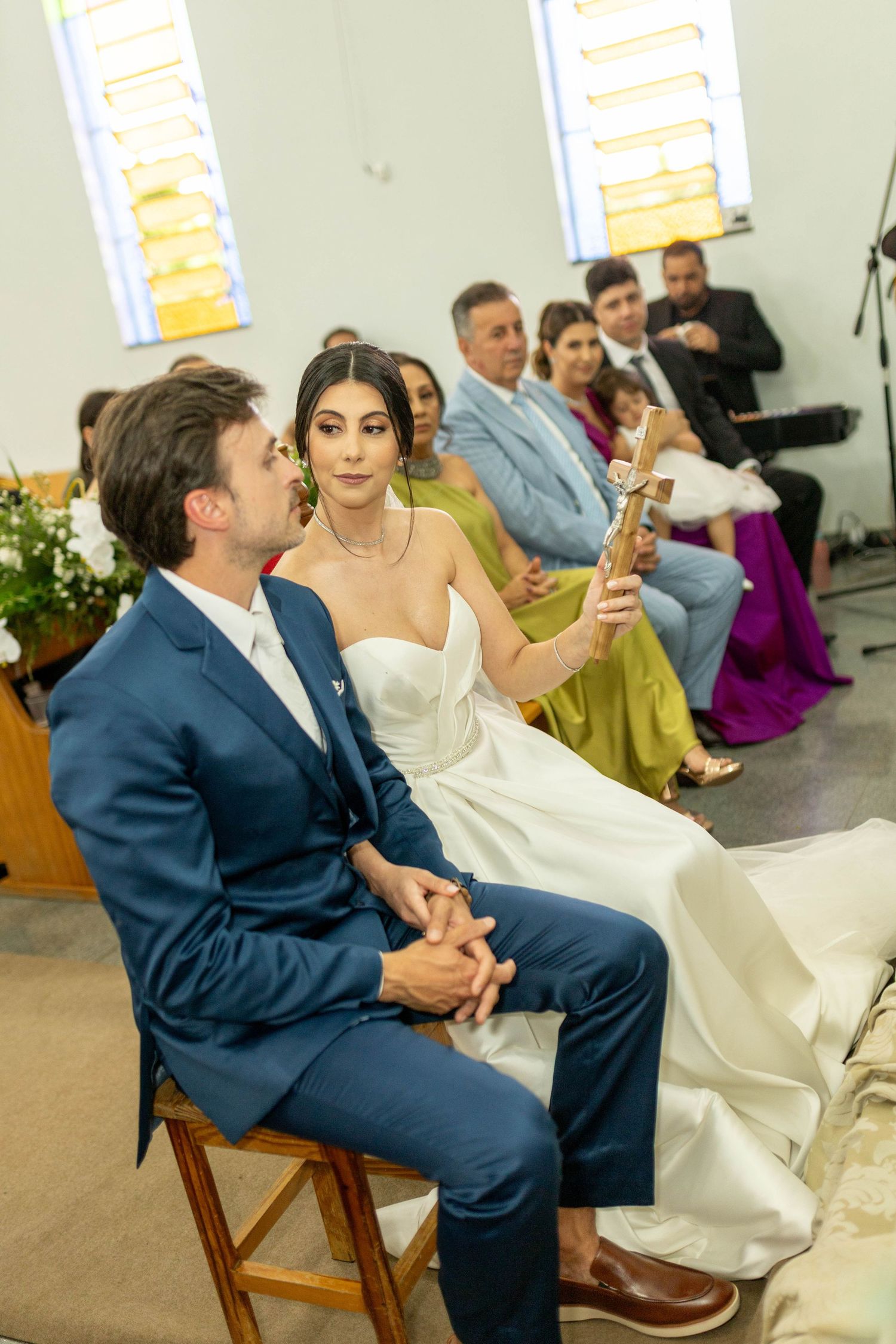 A bride in a white gown and groom in a blue suit sit together during their wedding ceremony in a church.