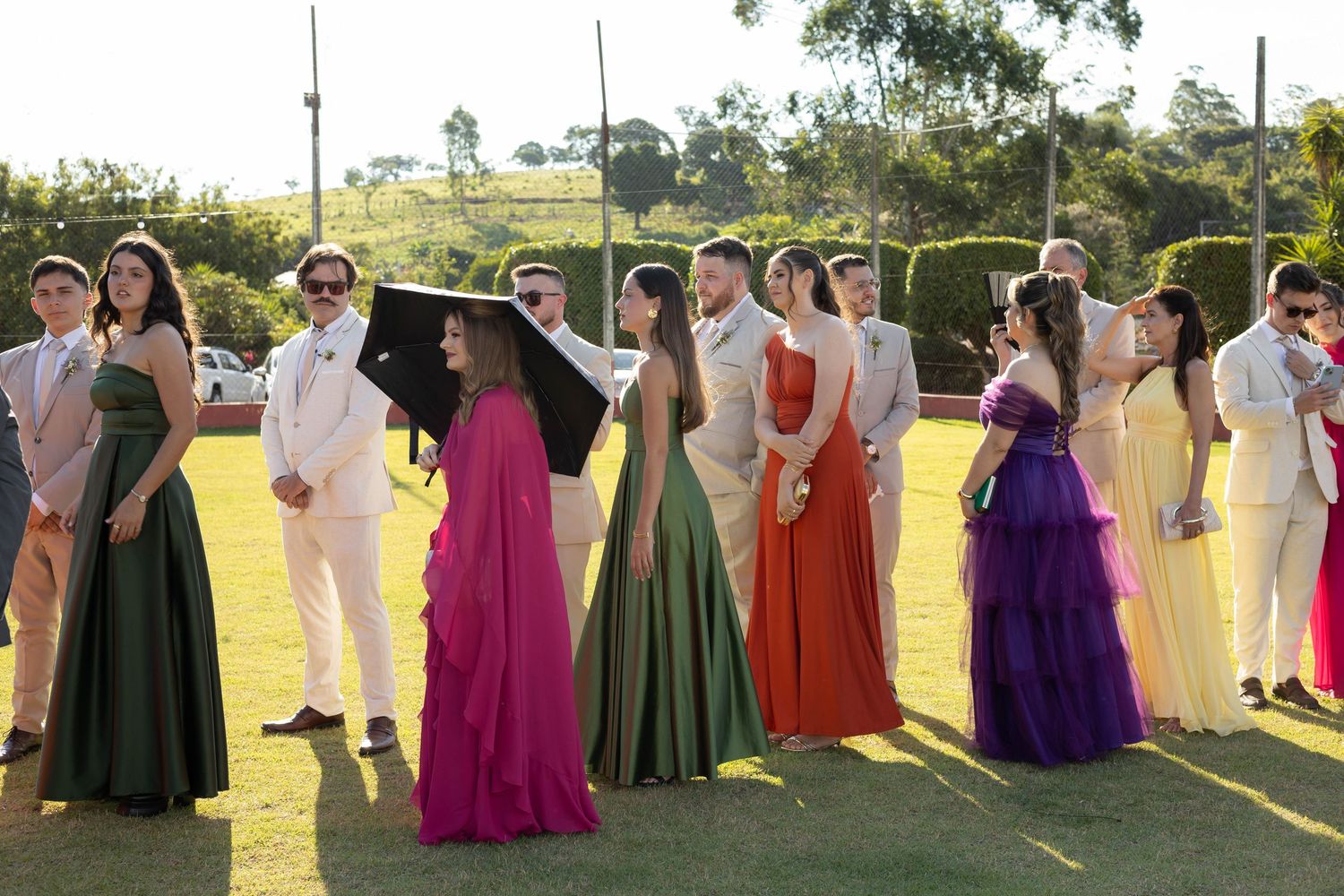 Group of teens in colorful formal gowns and suits posing outdoors on green grass at prom or formal event.
