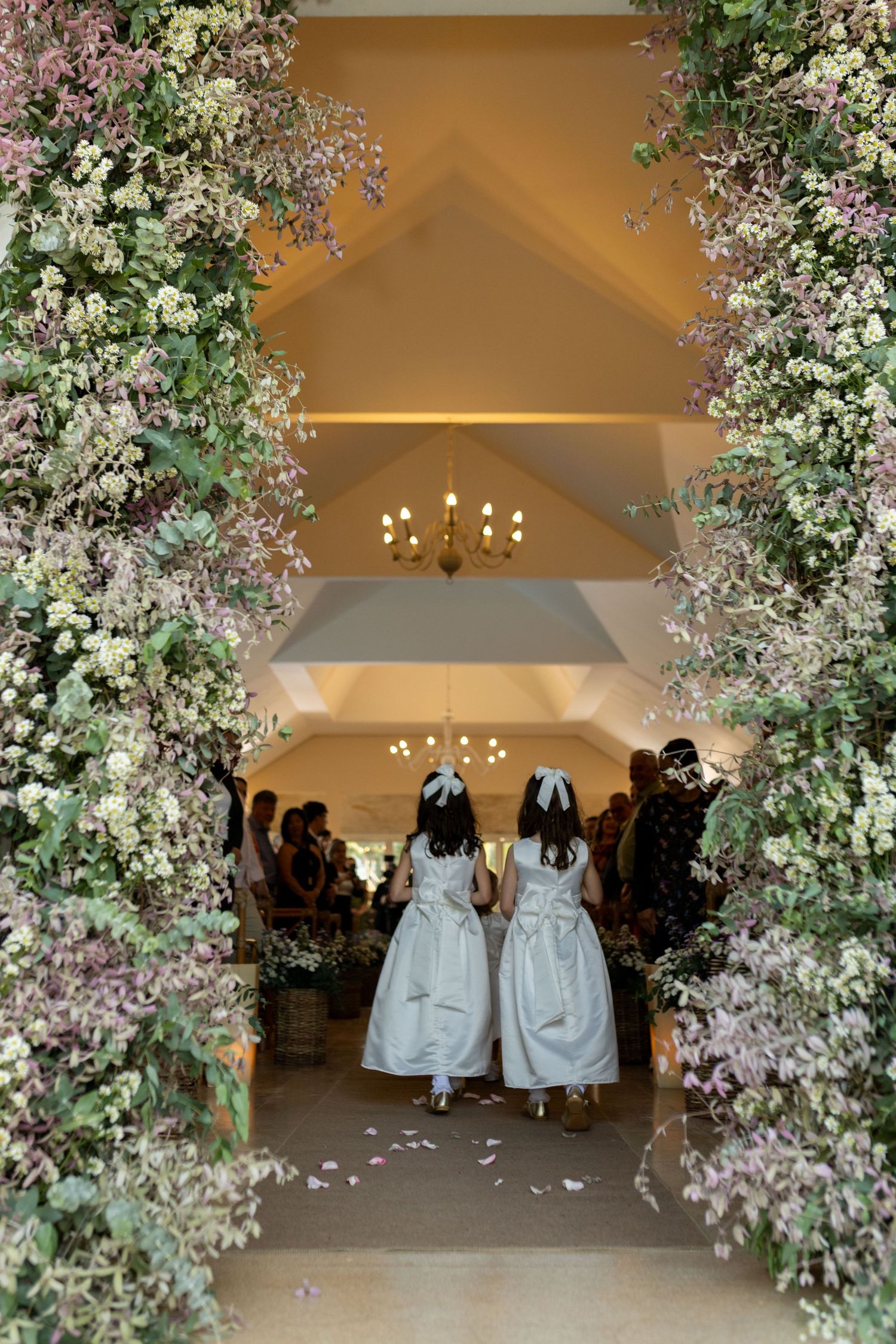 Two flower girls in white dresses walk through an elegant floral archway into a wedding ceremony venue.