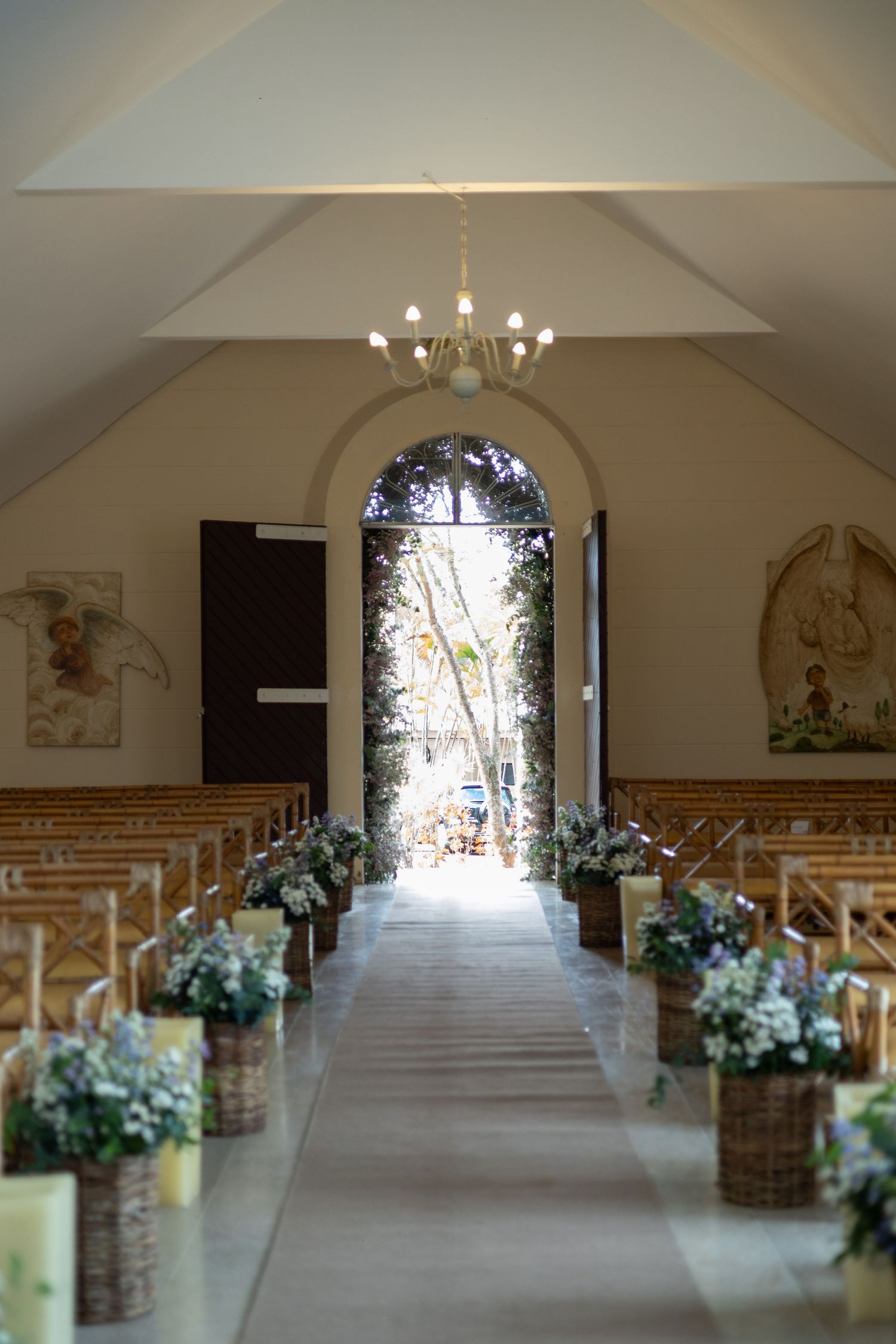 Elegant chapel wedding aisle with floral basket arrangements, wooden pews, chandelier, and arched doorway leading to sunlit garden.