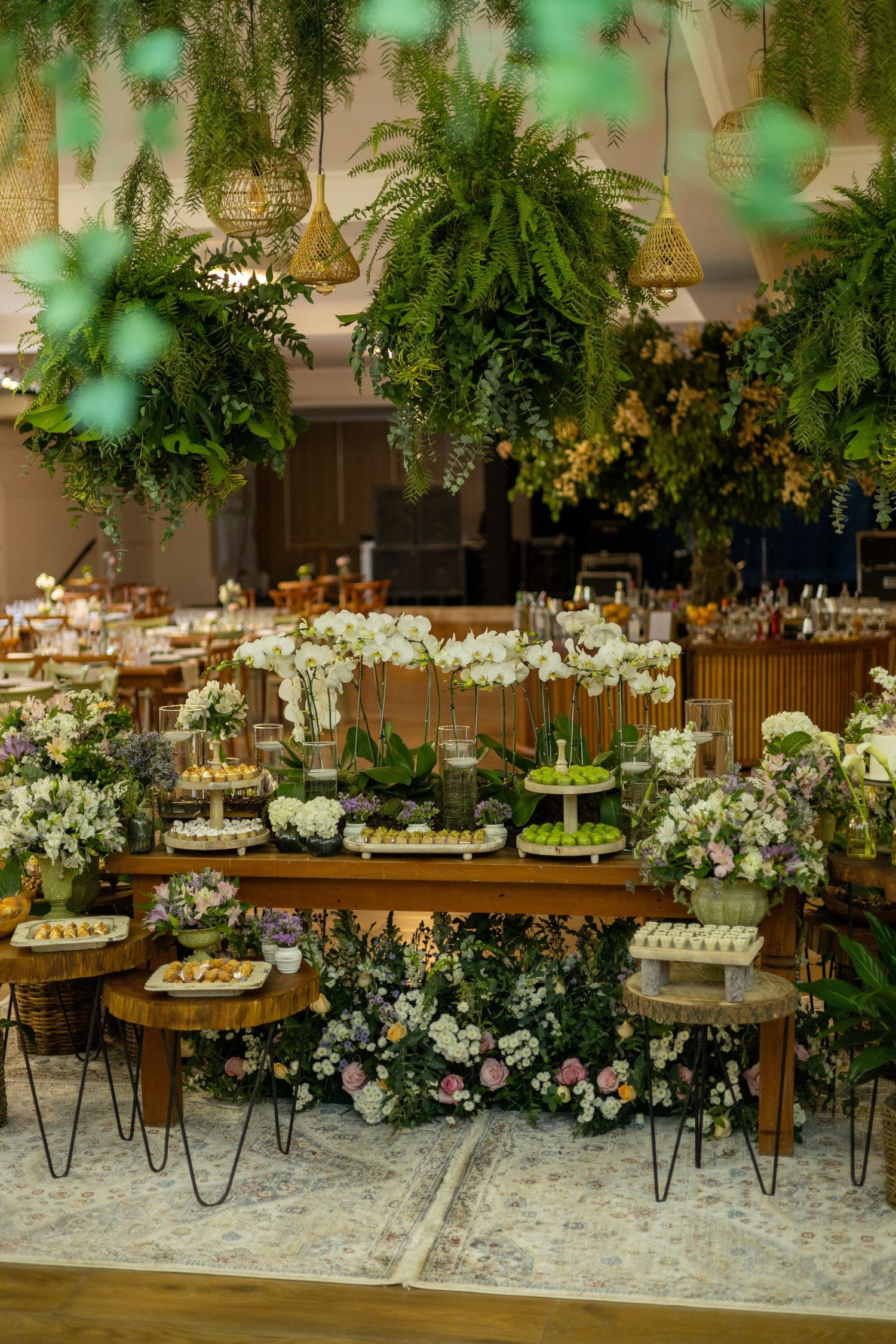 Elegant wedding dessert table adorned with lush greenery, white orchids, and hanging ferns in a beautifully decorated reception hall.