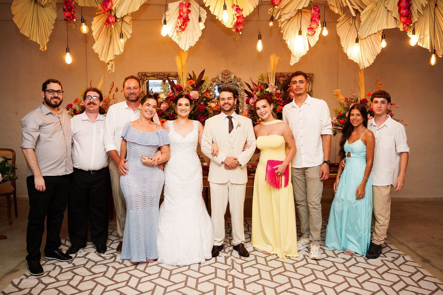 Wedding party group photo with bride in white gown and groom in ivory suit surrounded by guests under floral decor.