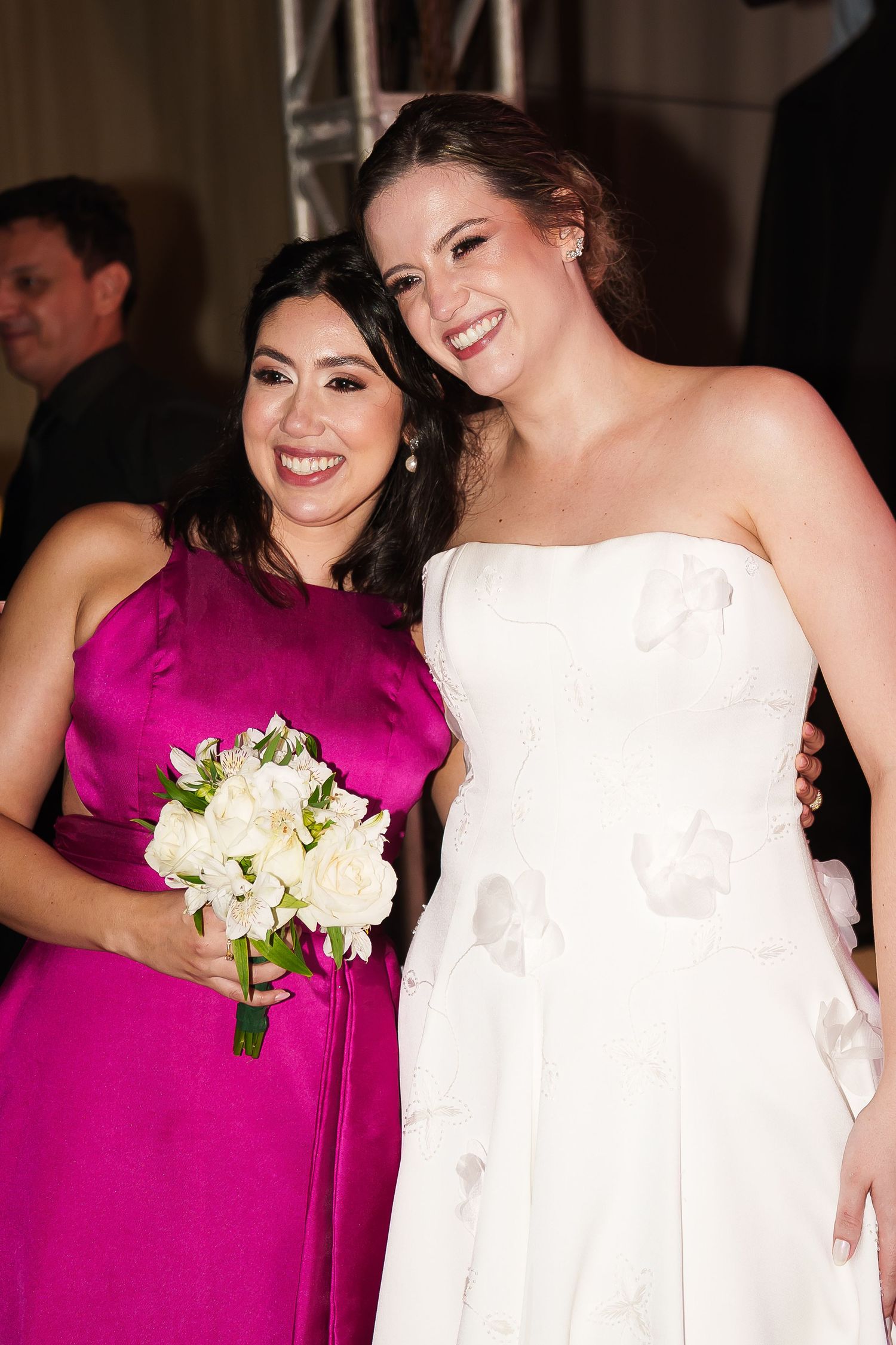 Bride in white strapless gown poses with bridesmaid in fuchsia dress holding white bouquet at wedding reception.