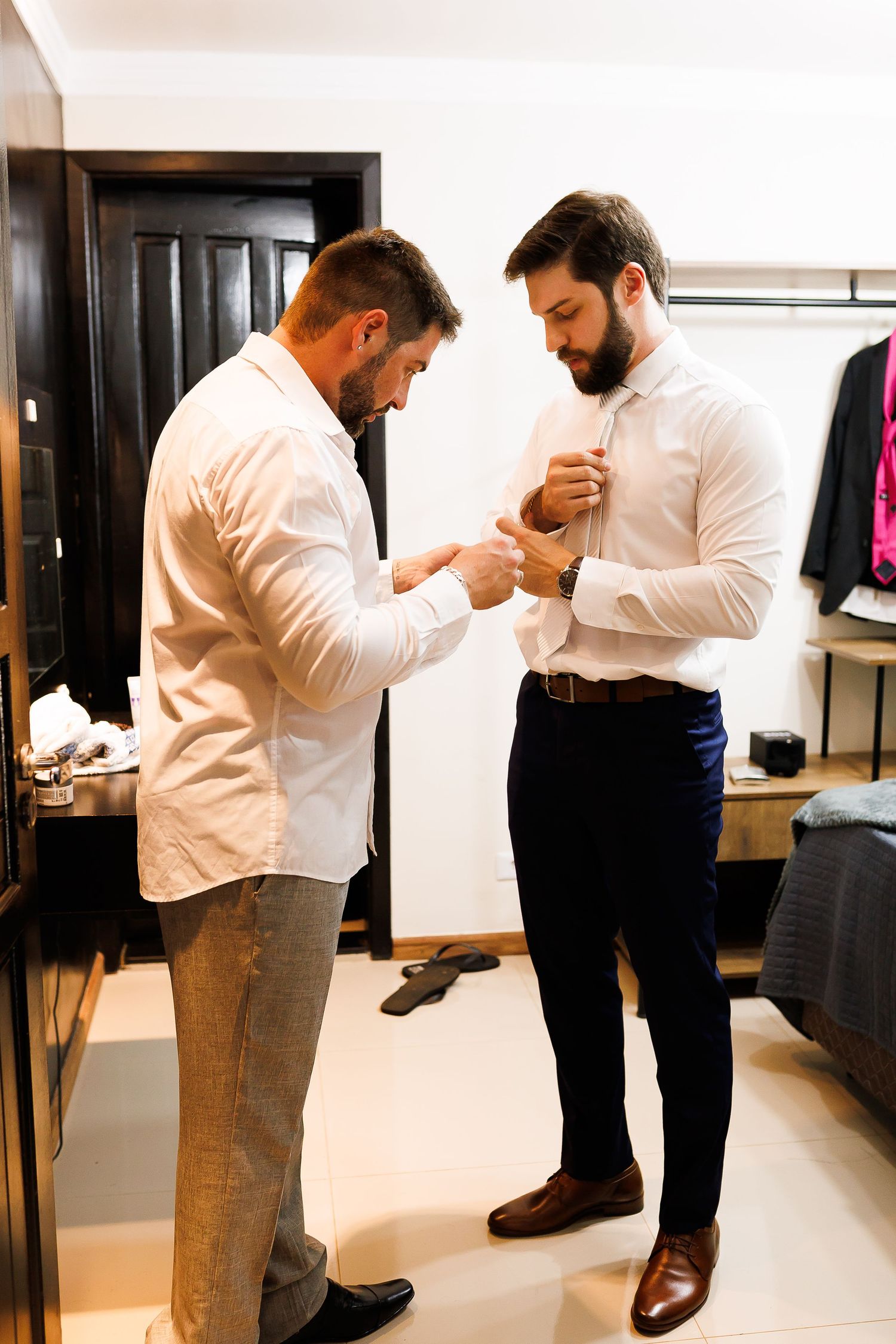 Two men in white shirts helping each other with cufflinks while getting dressed in a modern room.