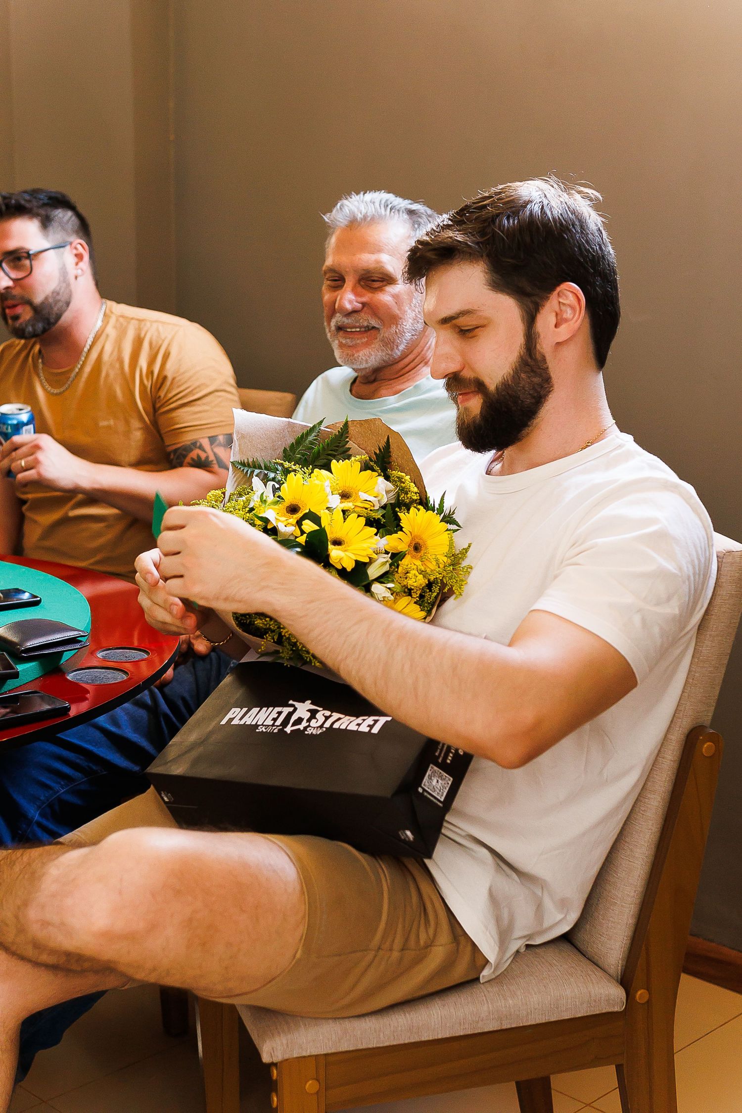 A bearded man in a white t-shirt holds yellow flowers and a Planet Street box, with two men smiling in the background.
