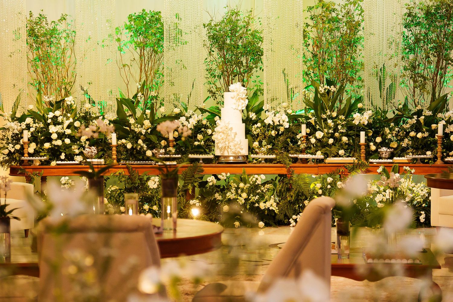 Elegant wedding reception with white tiered cake surrounded by lush greenery and white floral arrangements on a mirrored table.