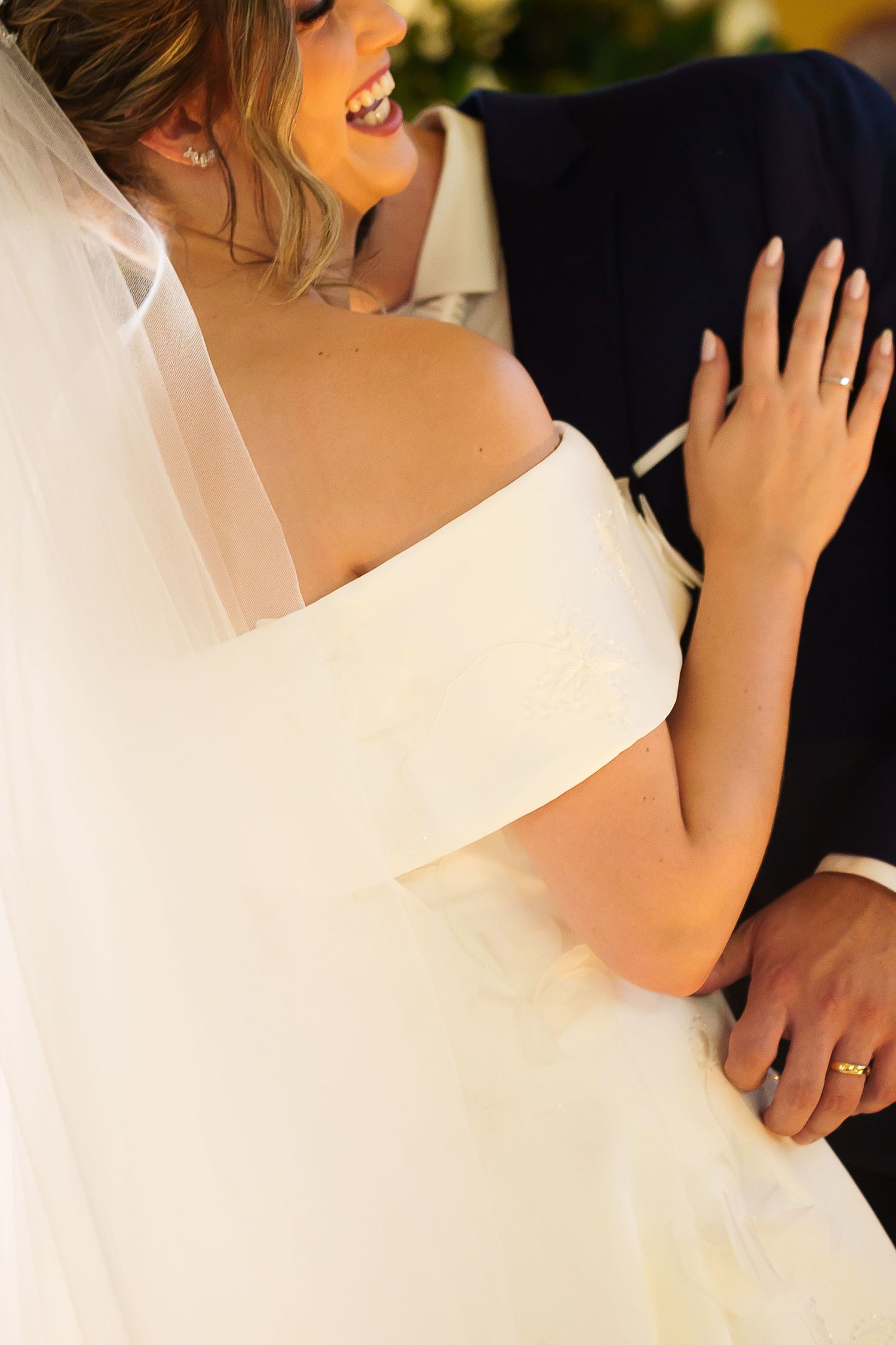 Bride in off-shoulder white gown and veil laughing with groom in navy suit at elegant wedding ceremony.