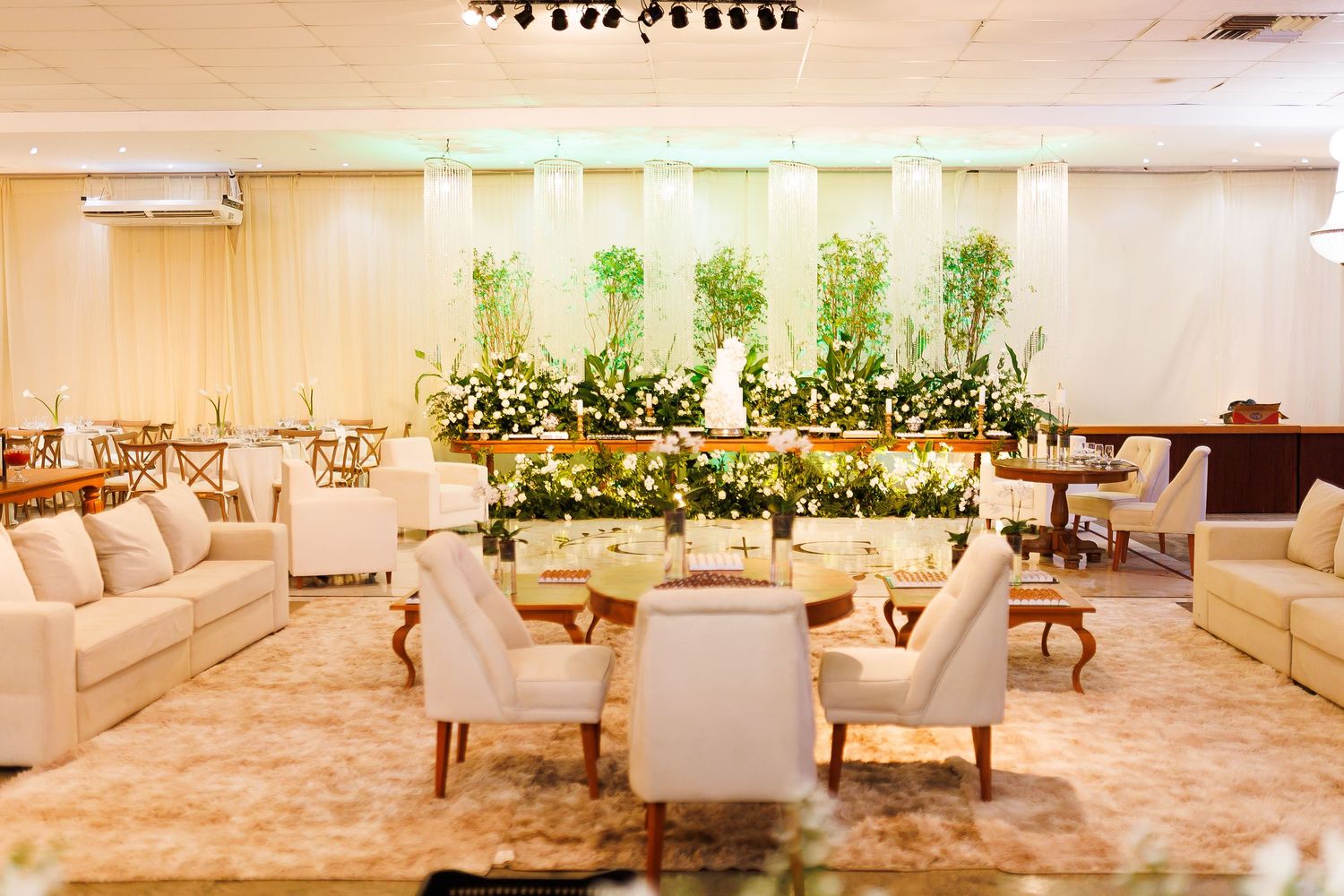 Elegant wedding reception hall with white sofas, floral arrangements, and lush greenery backdrop in a bright, airy space.