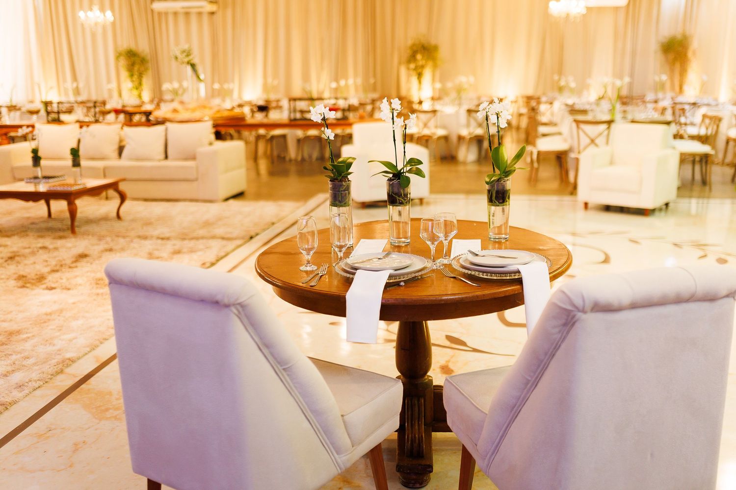 Elegant banquet hall with round wooden tables, white chairs, and floral centerpieces in a warmly lit reception venue.