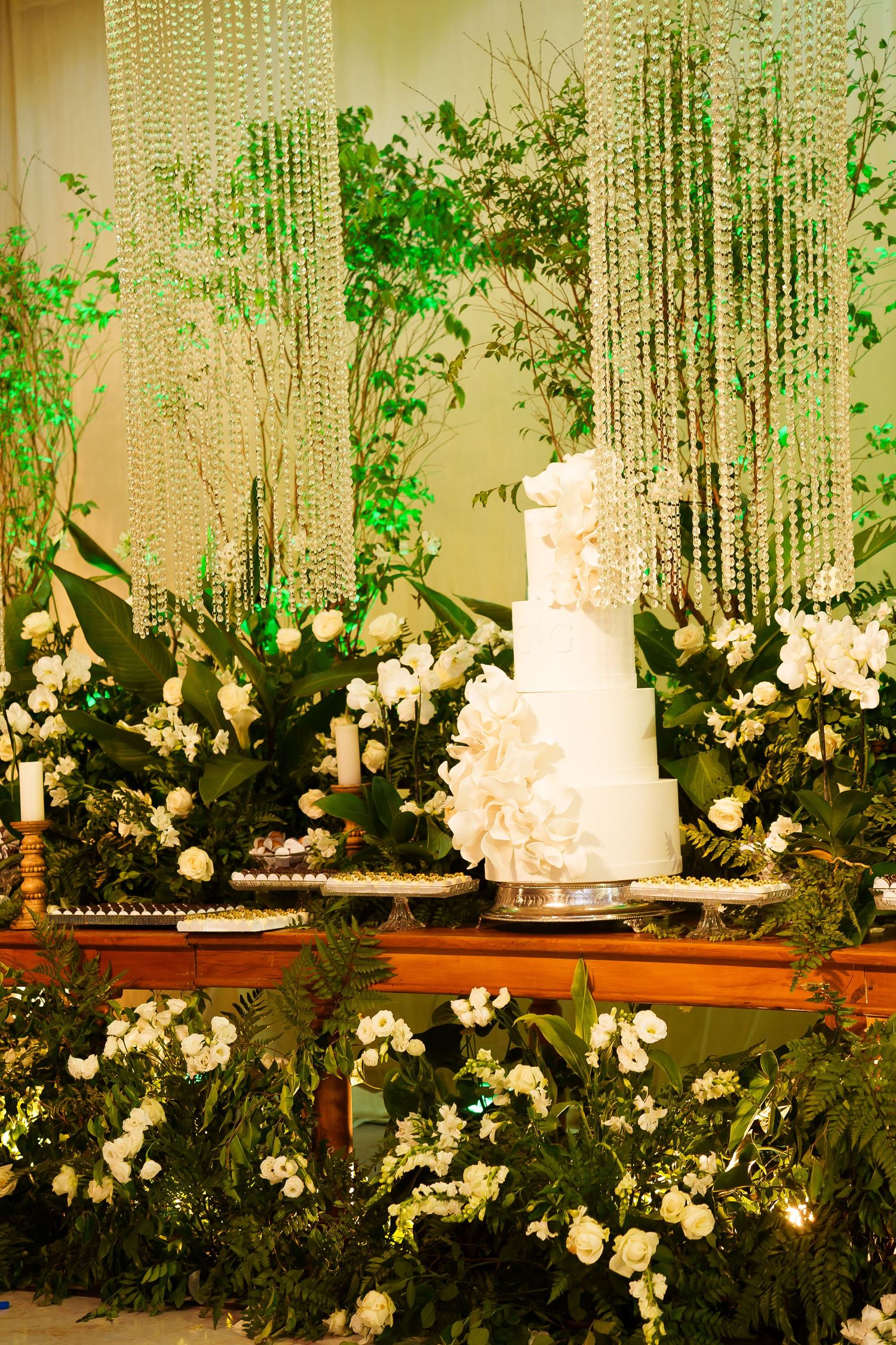 Elegant white tiered wedding cake surrounded by lush greenery, white flowers, and cascading crystal beads on a decorated table.
