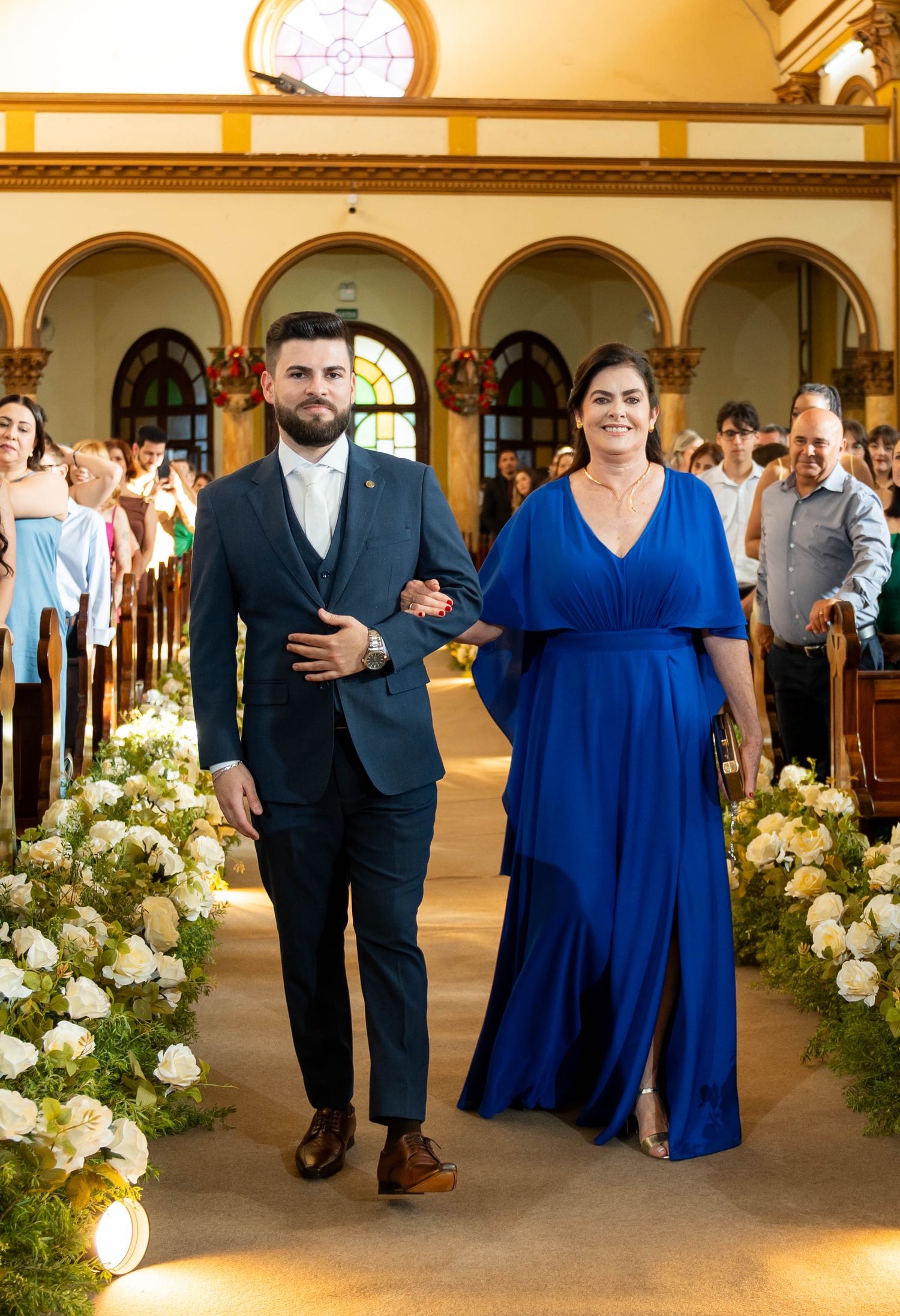 A man in a navy suit escorts a woman in a royal blue gown down a flower-lined church aisle at a wedding.
