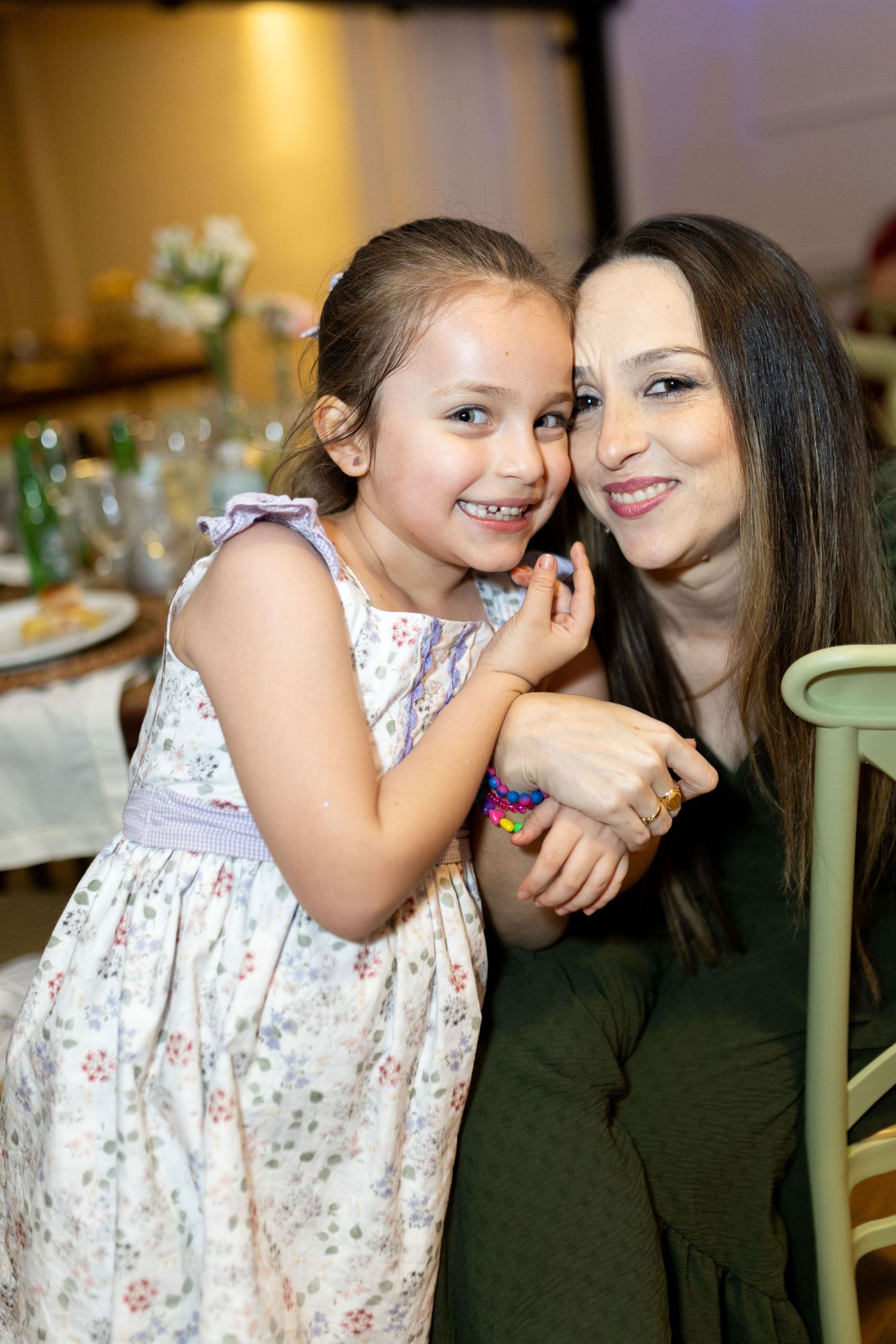 A young girl in a floral dress smiles while hugging a dark-haired woman at a restaurant table.