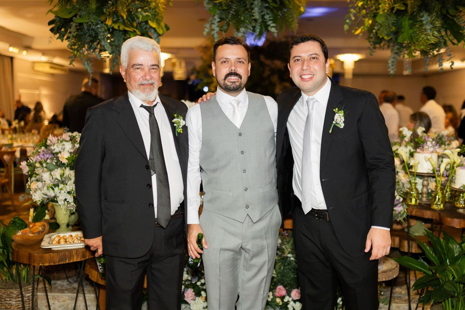 Three men in formal attire pose together at an elegant wedding reception with floral decorations.