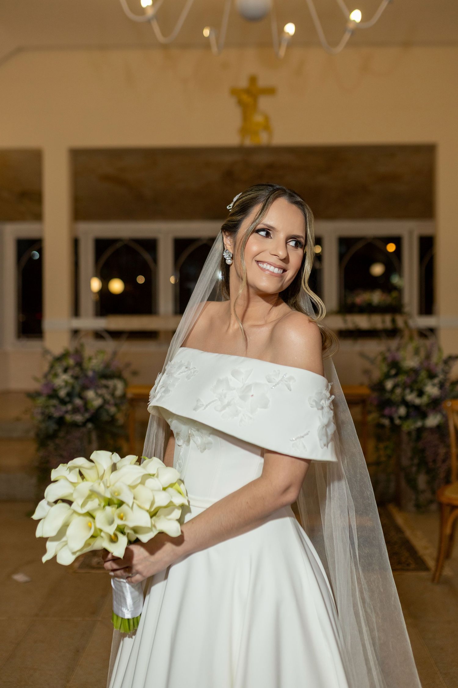 Smiling bride in off-shoulder white gown holds calla lily bouquet inside decorated church with crucifix.