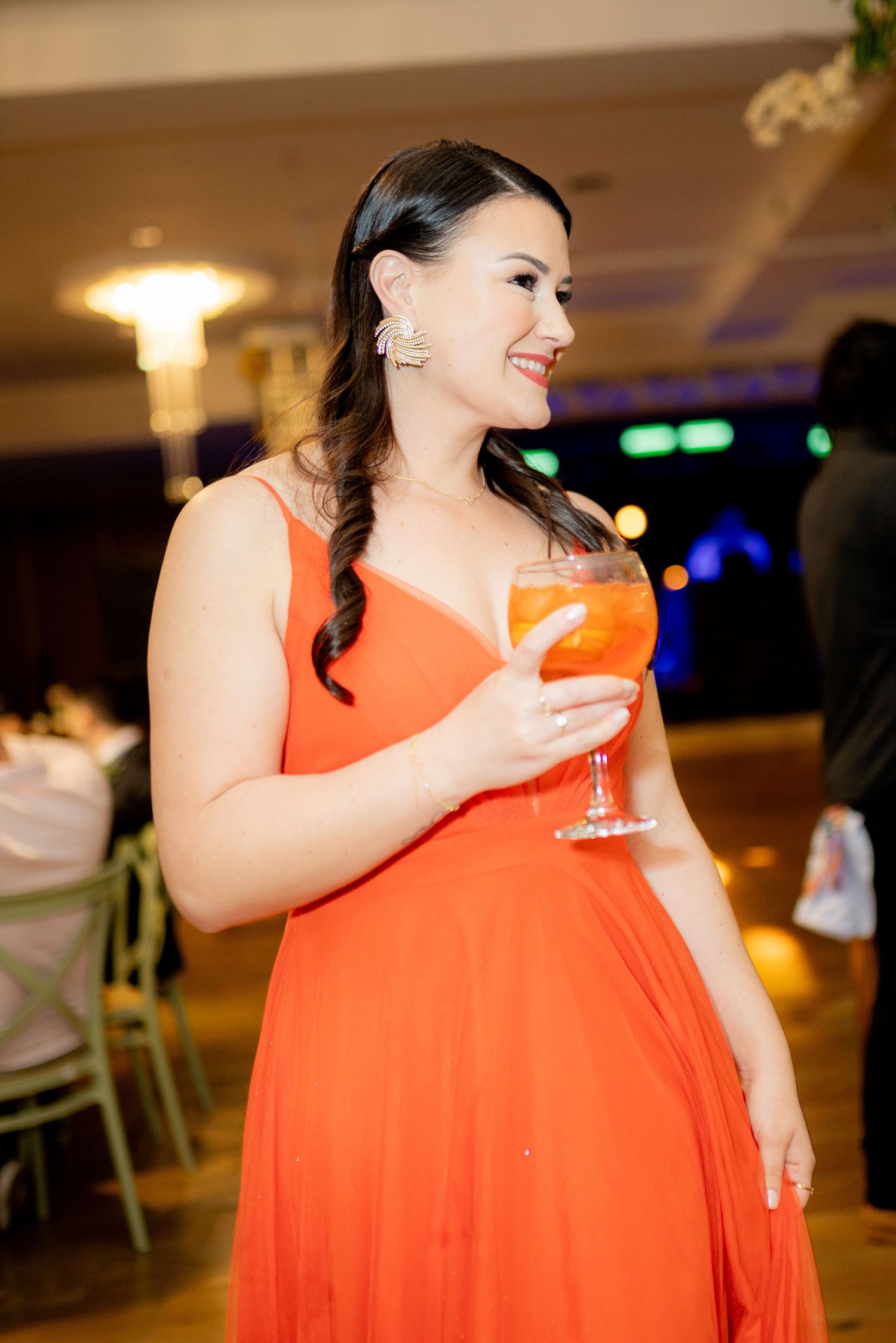 Woman in orange gown holding a cocktail at an elegant event, smiling and wearing gold earrings.