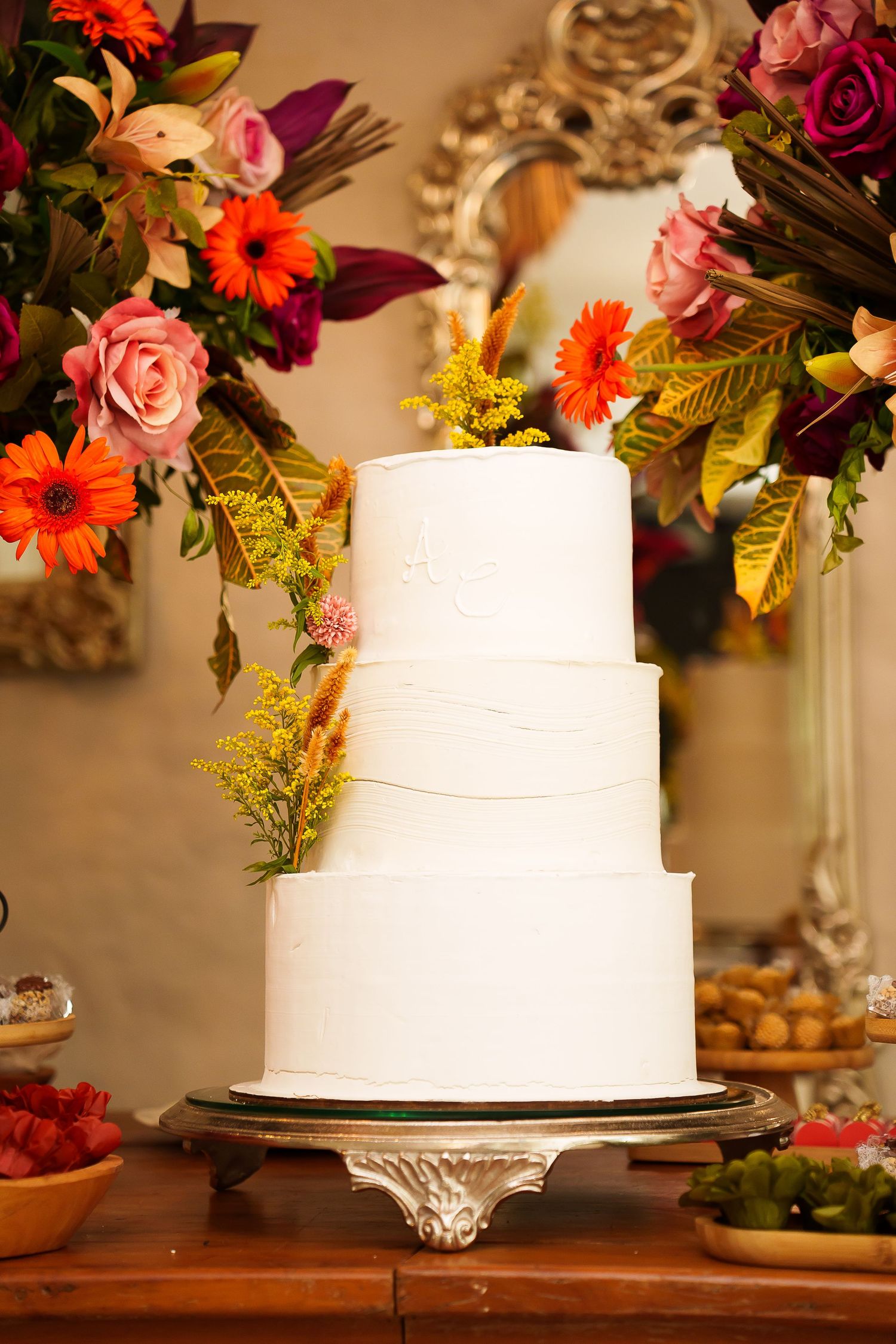 Elegant three-tier white wedding cake adorned with yellow flowers, displayed on a silver stand surrounded by colorful floral arrangements.