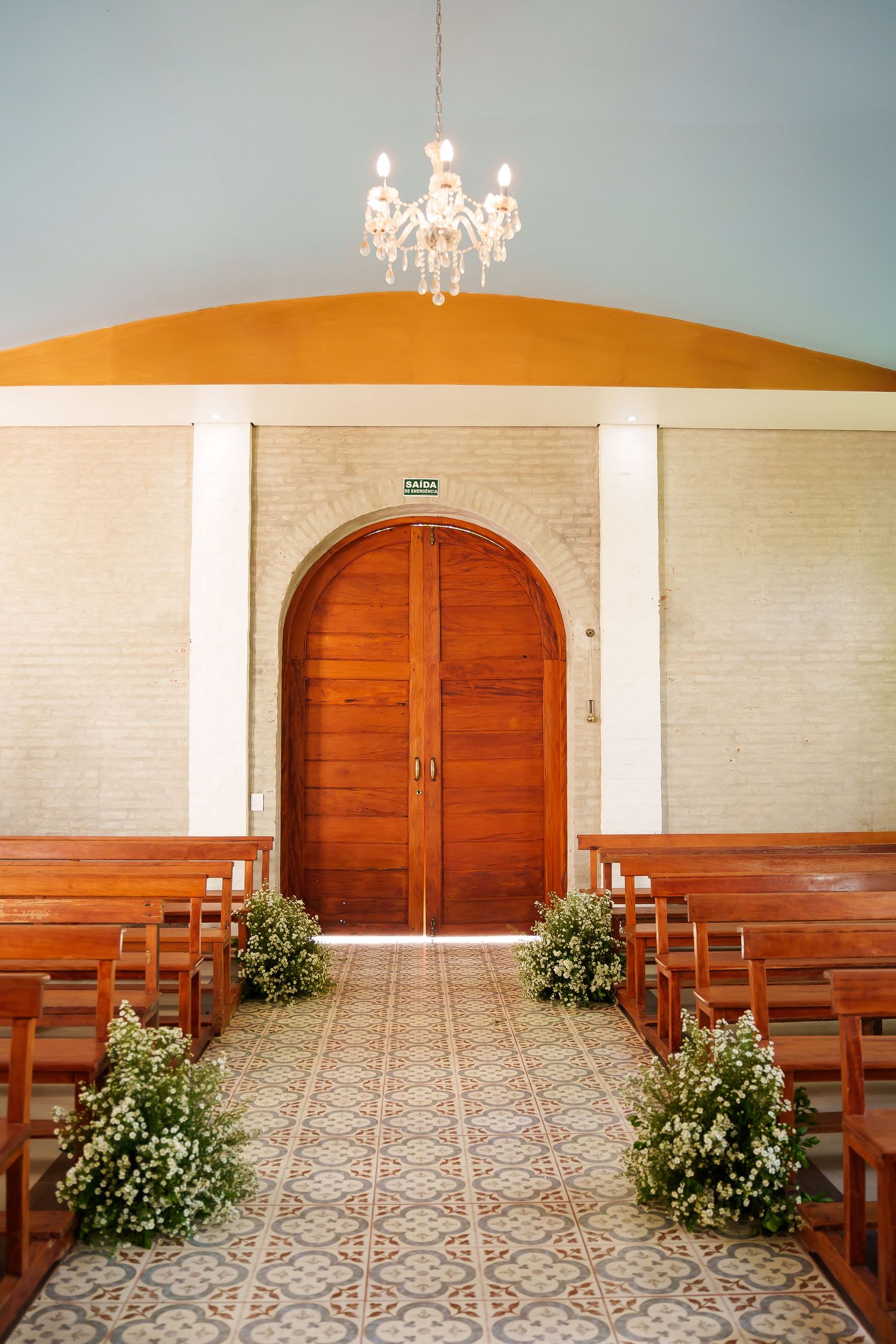 Elegant chapel interior with wooden pews, floral arrangements, arched door, chandelier, and decorative tile floor.