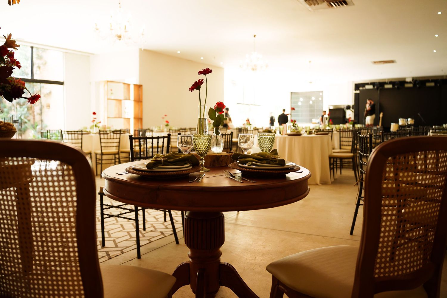 Elegant restaurant dining room with round wooden table, cane chairs, floral centerpiece, and softly lit banquet tables in background.