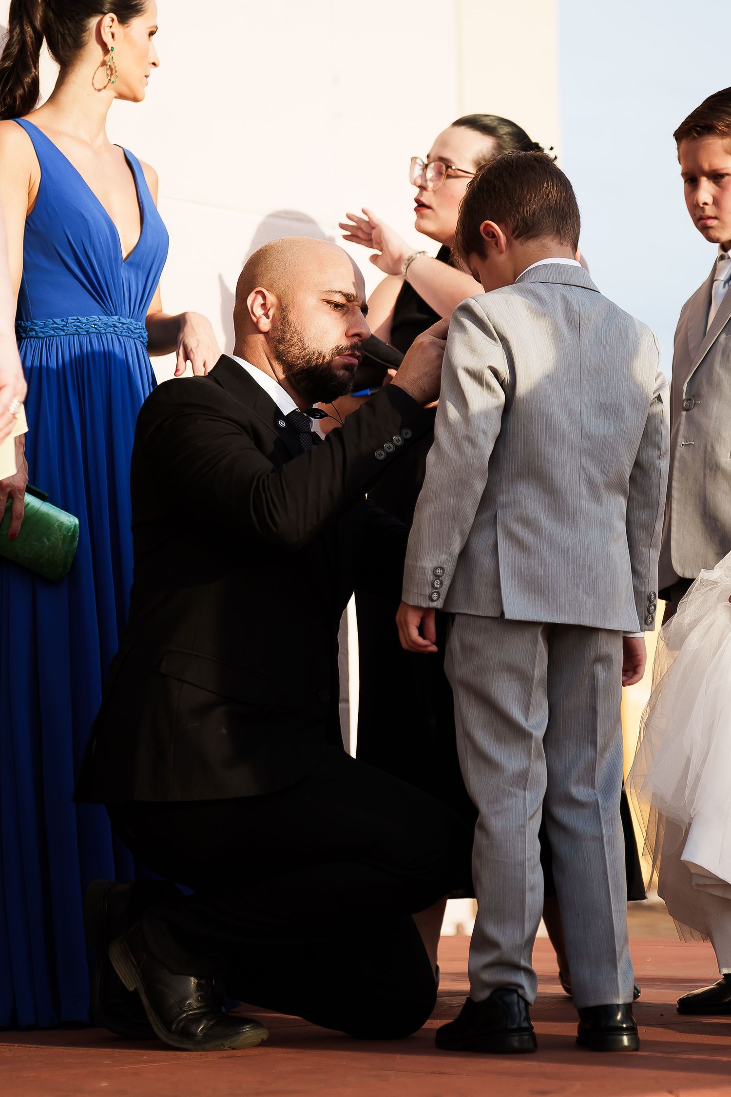 Bald man in black suit kneeling to adjust boy's gray suit jacket at formal event with elegantly dressed guests.