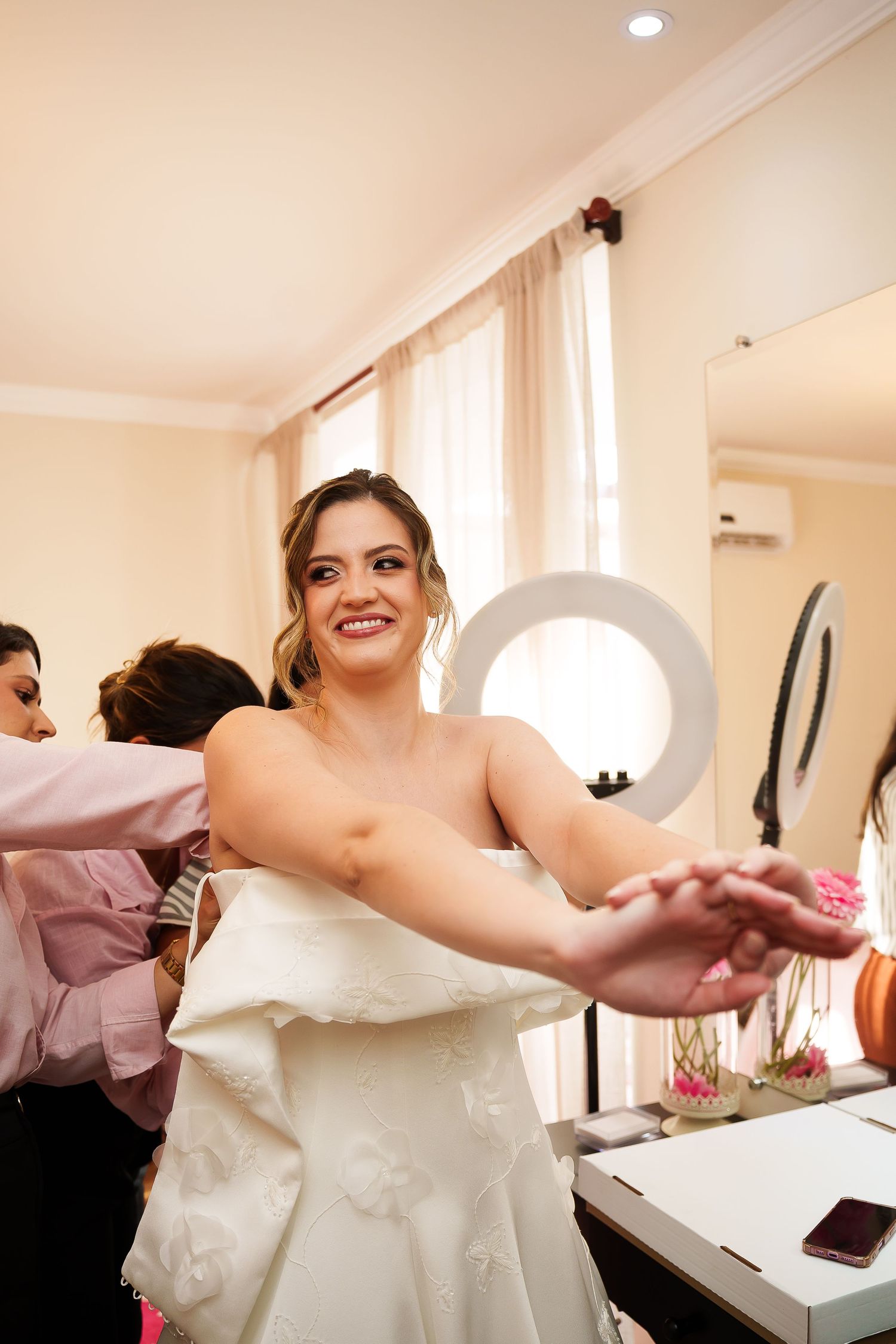 Smiling bride in white dress stretches arms out while getting ready, with ring light and mirror visible in background.