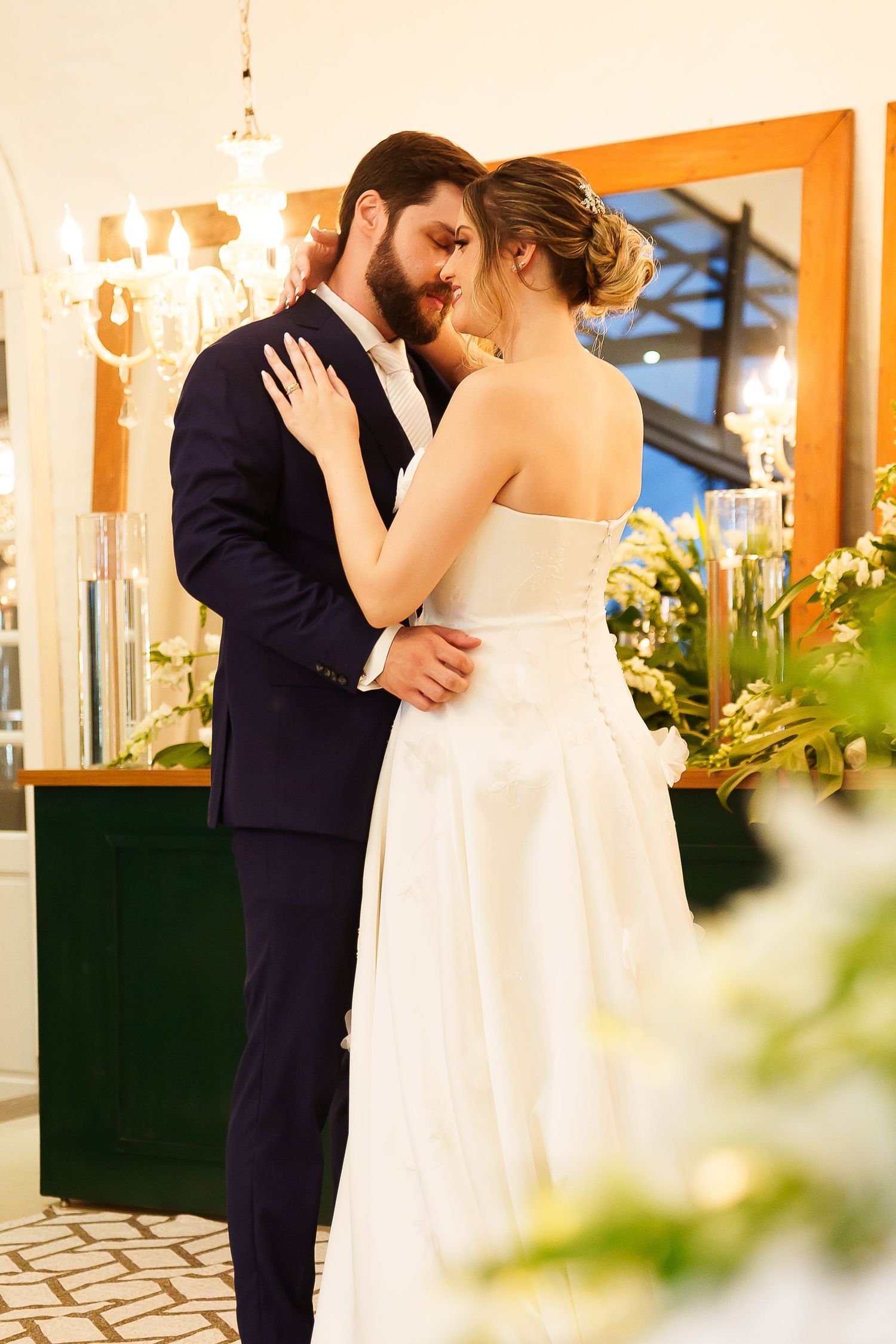 A bride and groom share a romantic embrace at their wedding reception, surrounded by floral arrangements and elegant decor.