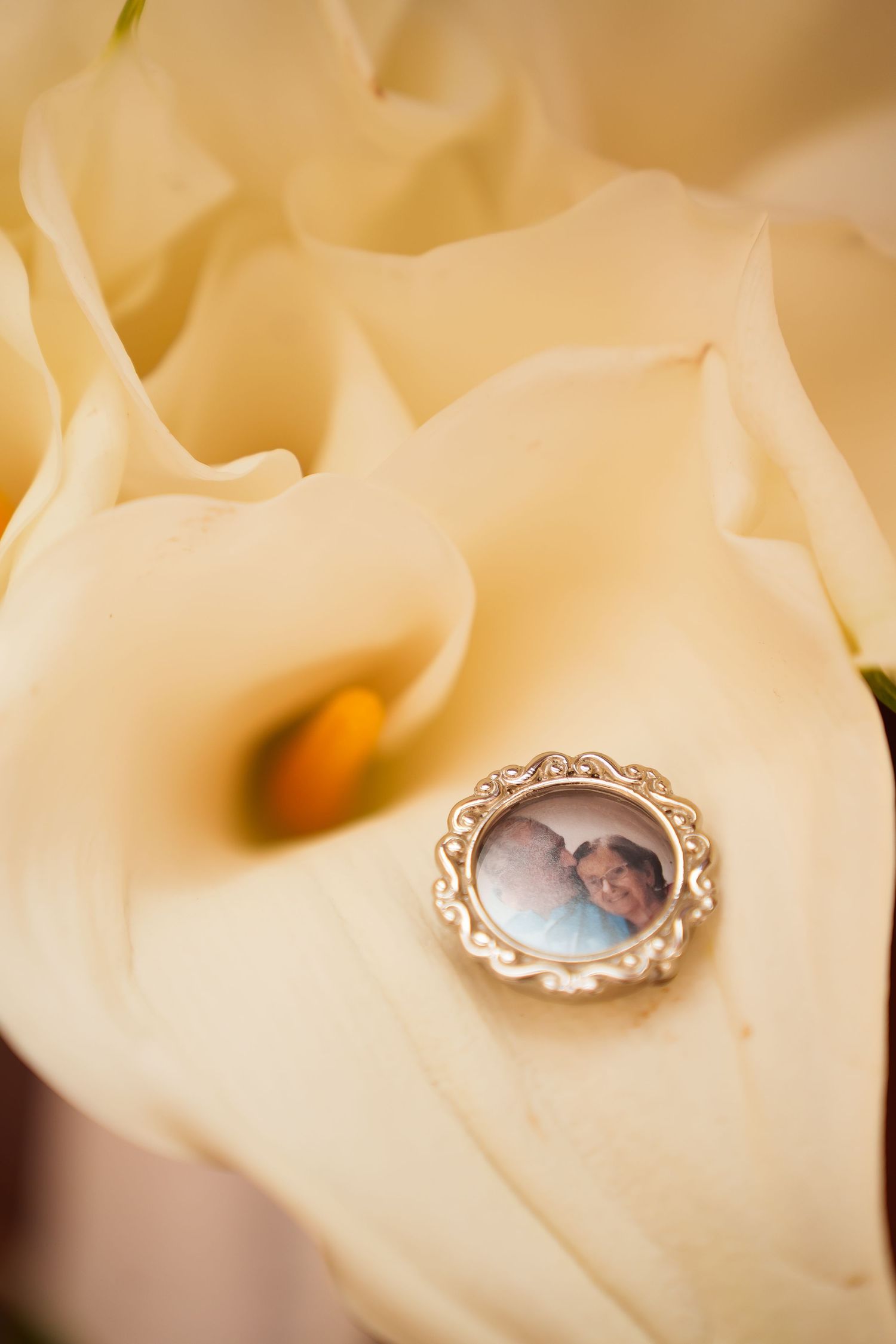 A silver locket with a photo resting on cream calla lily petals in a bridal bouquet.
