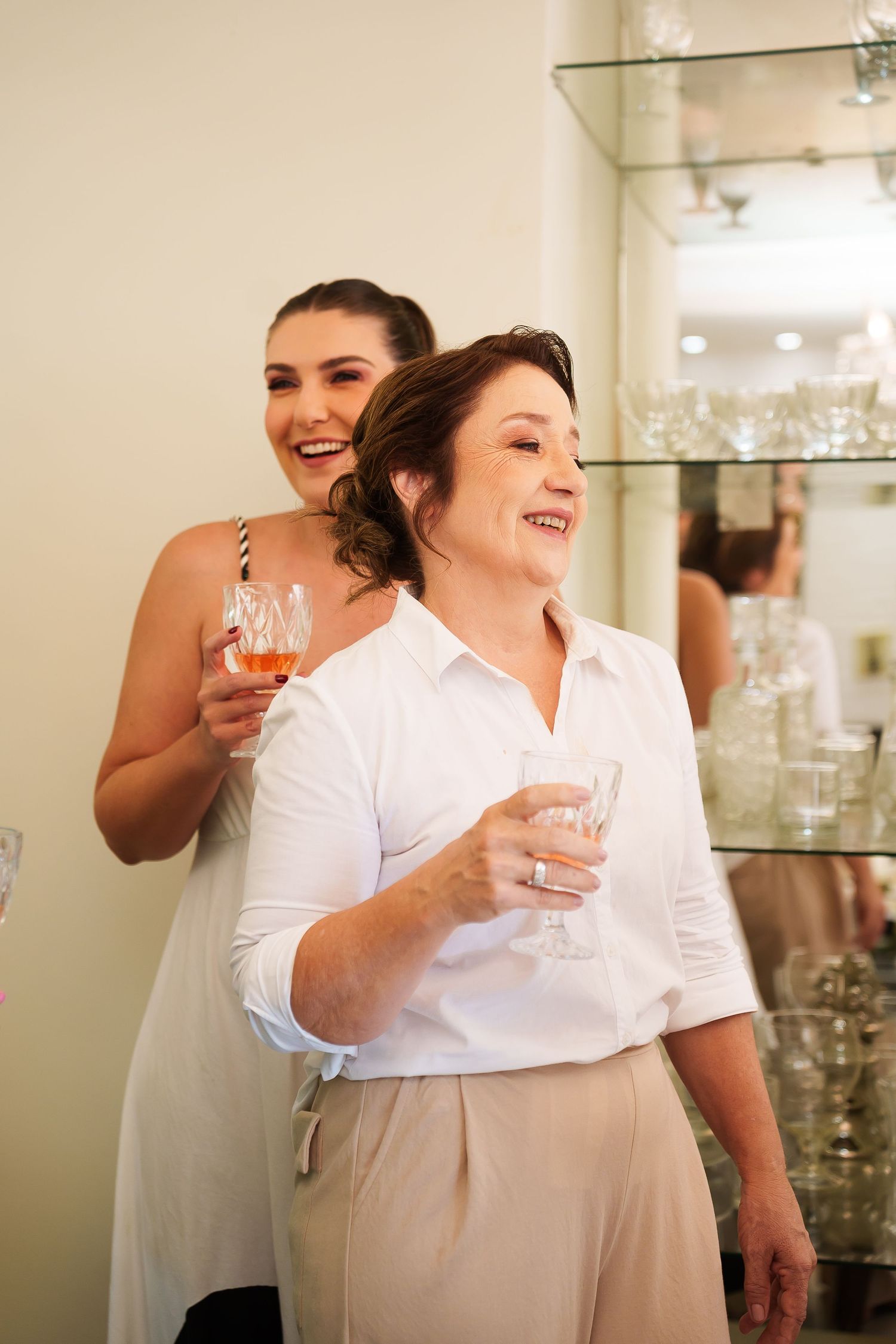 Two women smiling and holding drinks at a celebration, one in a white dress and the other in a white shirt.