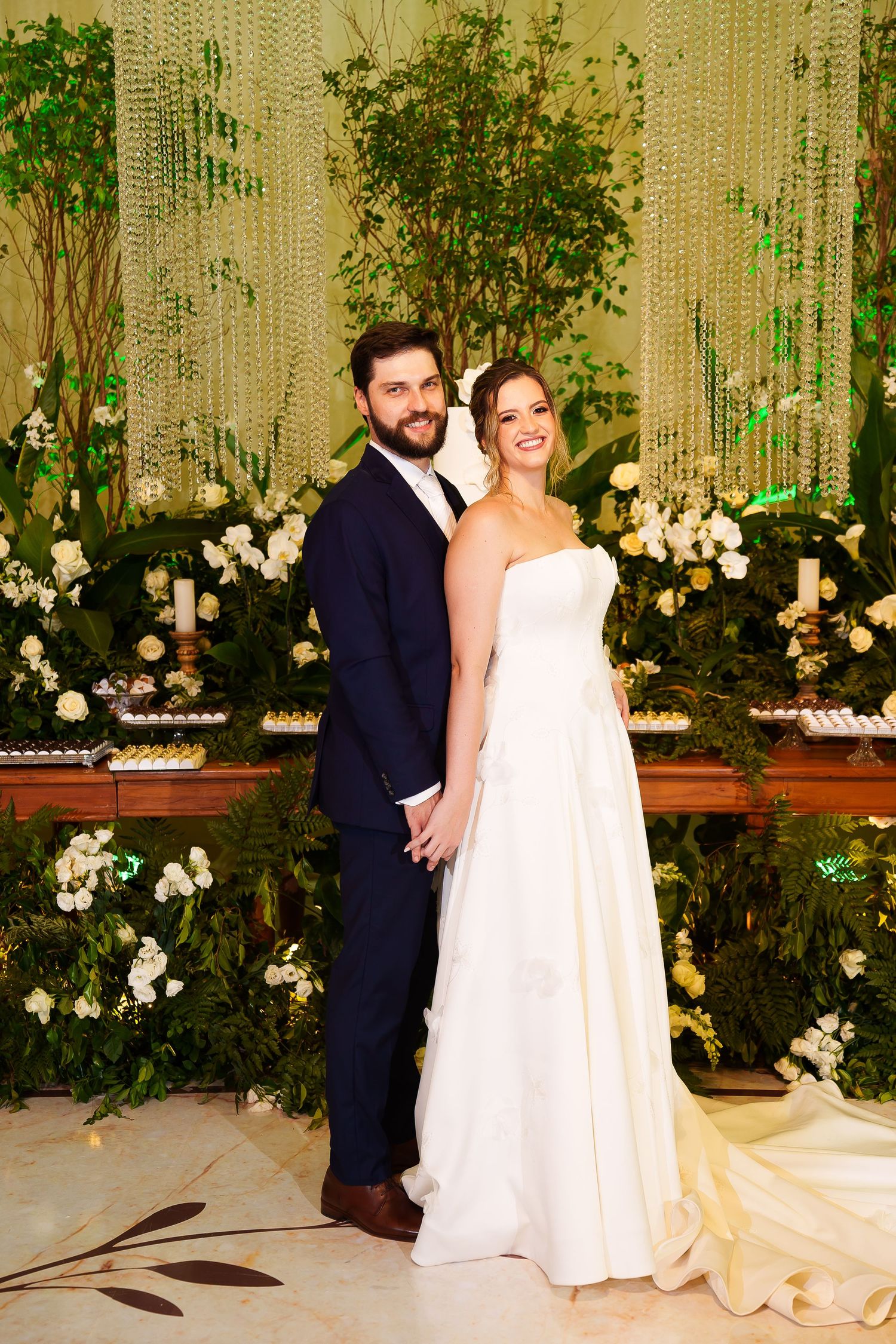 Bride in white strapless gown and groom in navy suit pose together at elegant wedding reception with floral backdrop.