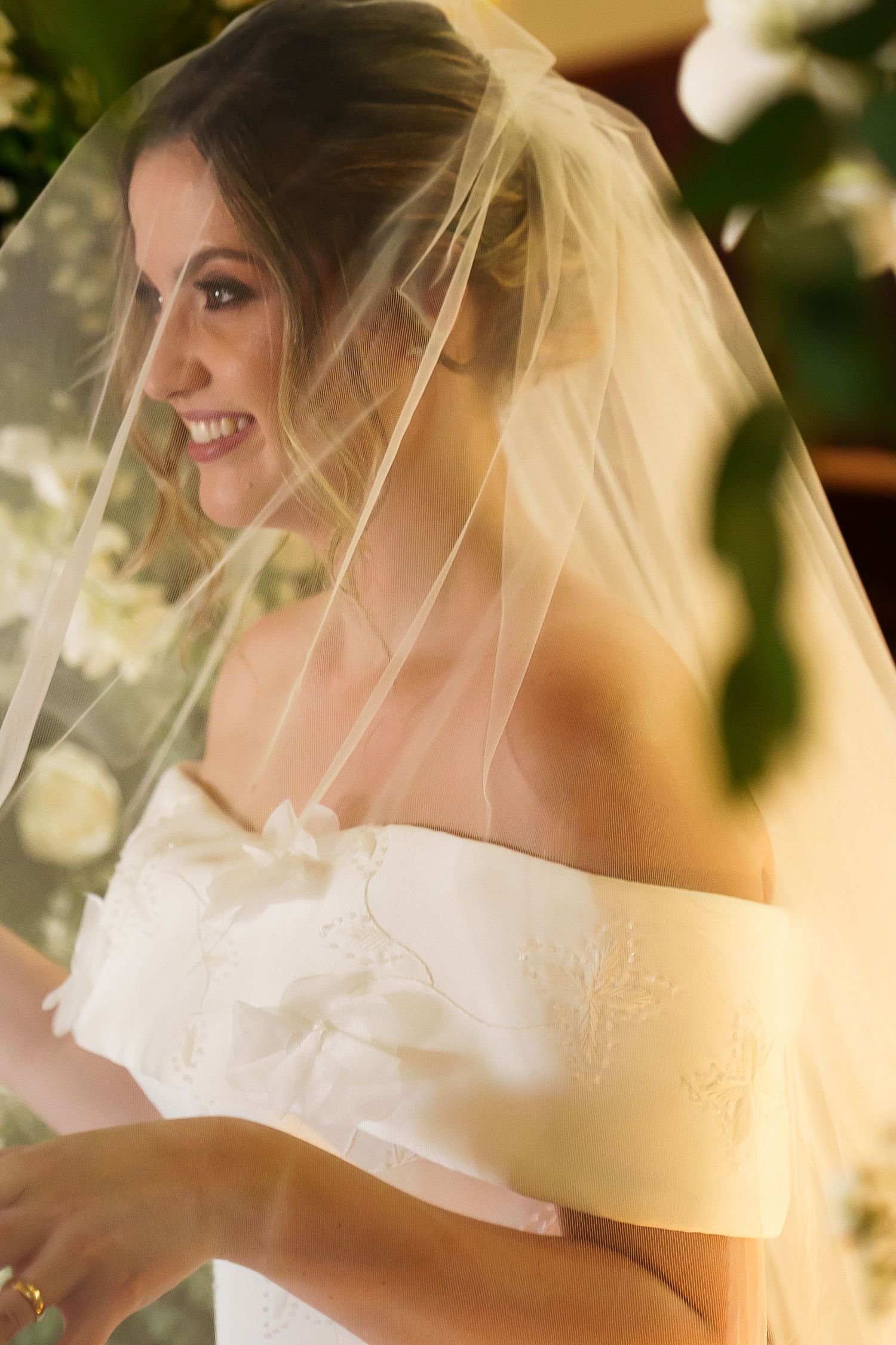 Smiling bride in off-shoulder white gown and sheer veil, surrounded by white flowers in warm light.