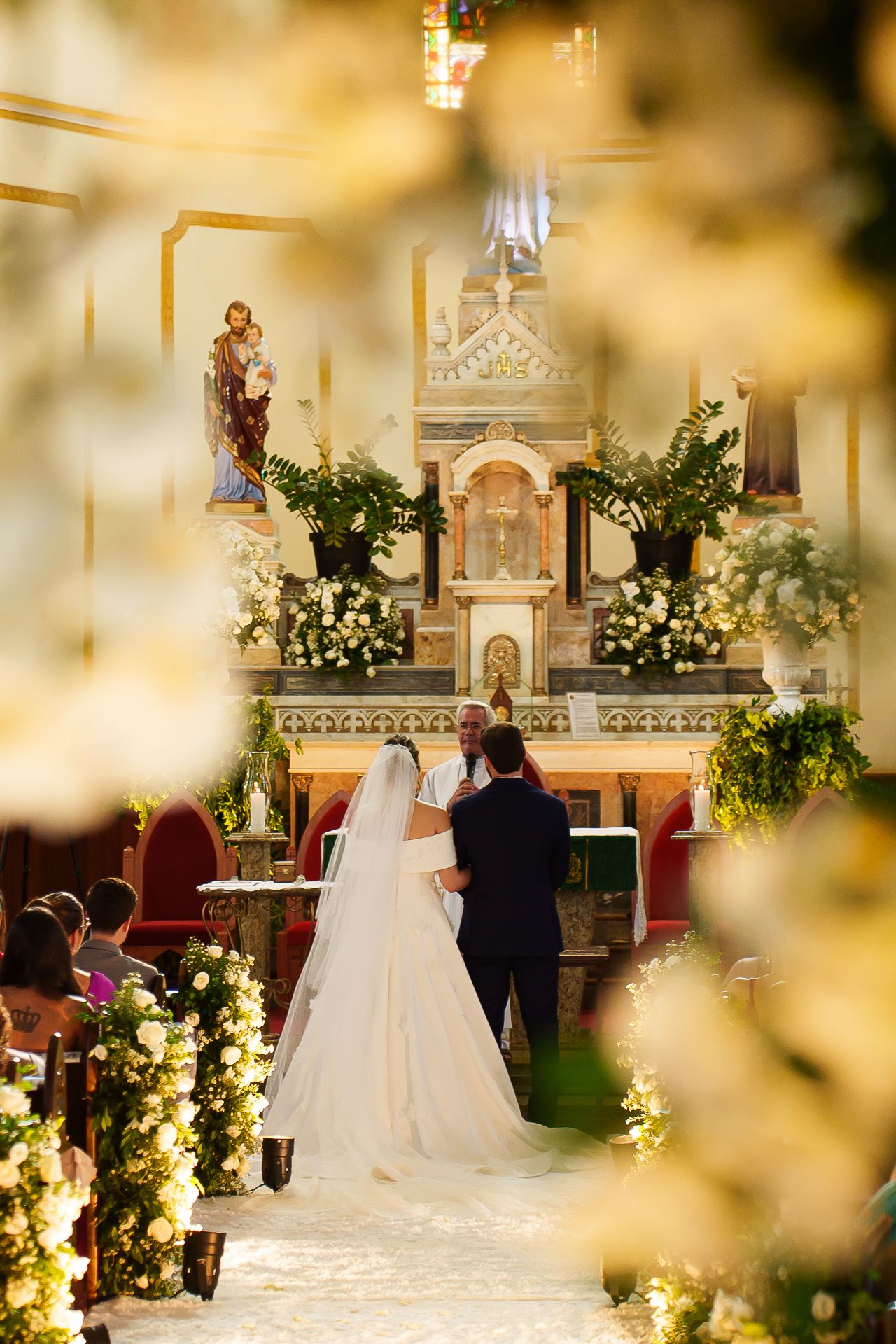 Bride and groom standing at ornate church altar during wedding ceremony, surrounded by lush white floral arrangements.