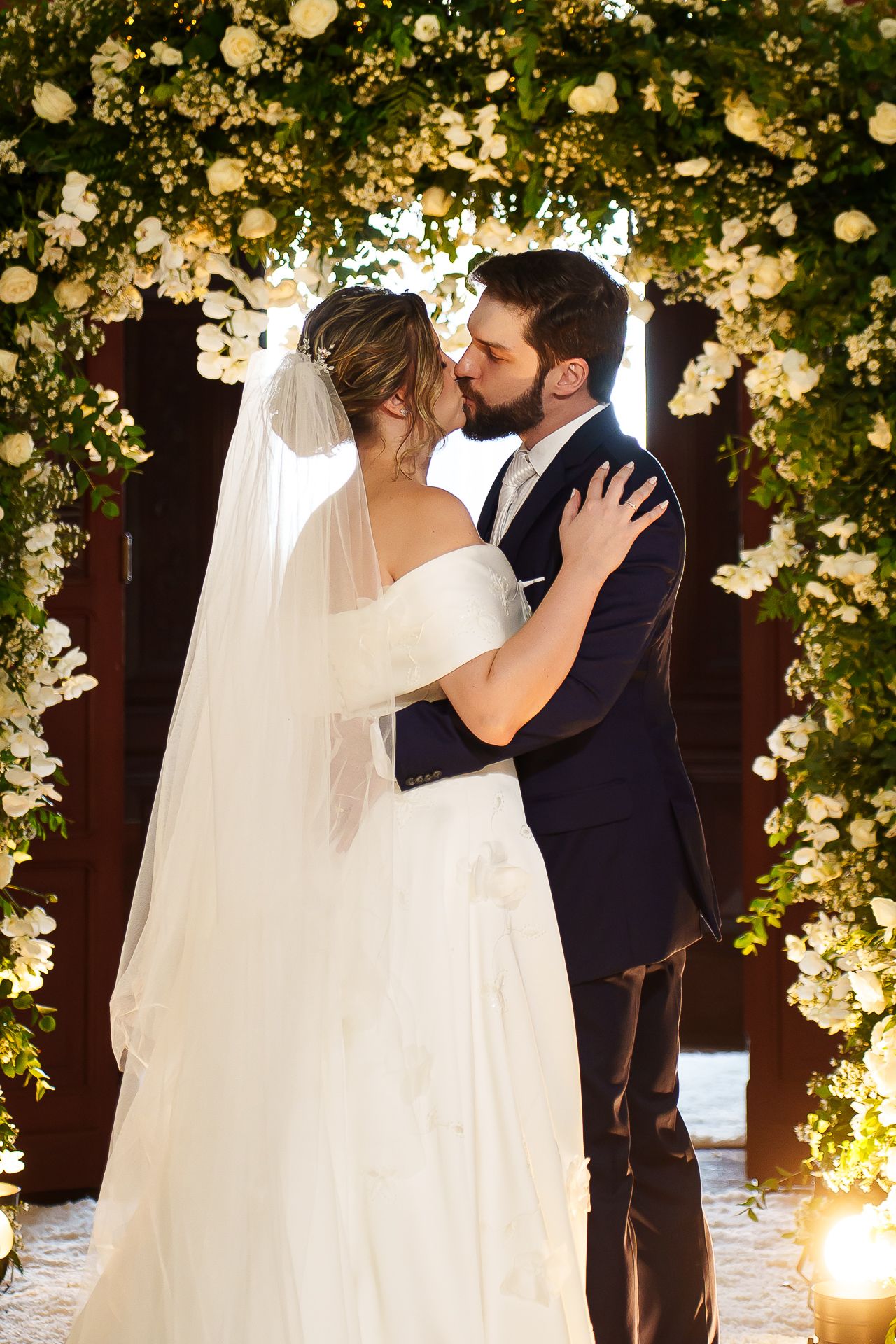 Bride and groom share a first kiss under a lush floral arch adorned with white roses at their wedding ceremony.