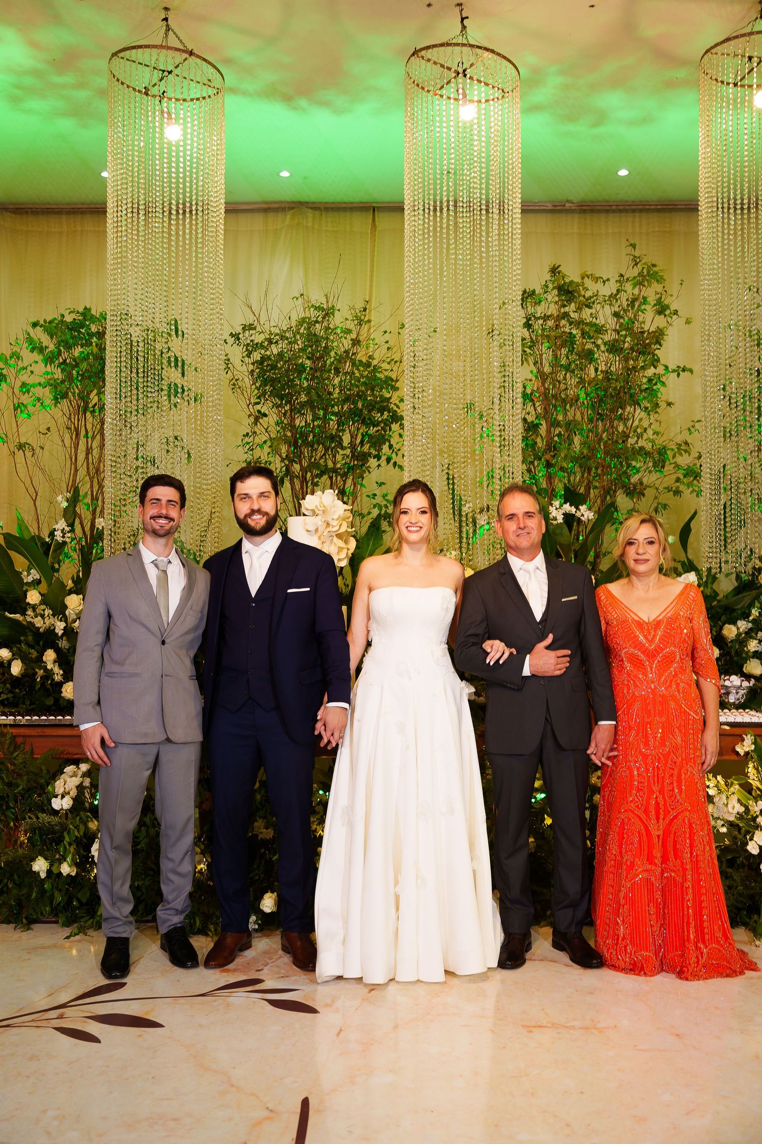 Wedding party of five posing elegantly in a decorated venue with crystal chandeliers and lush greenery backdrop.