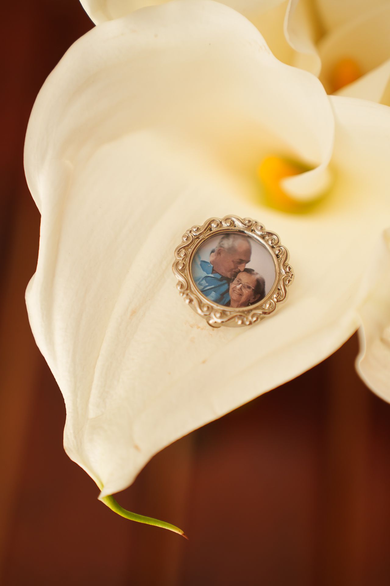 A silver photo brooch with a couple's portrait resting on a white calla lily petal, a sentimental wedding bouquet charm.