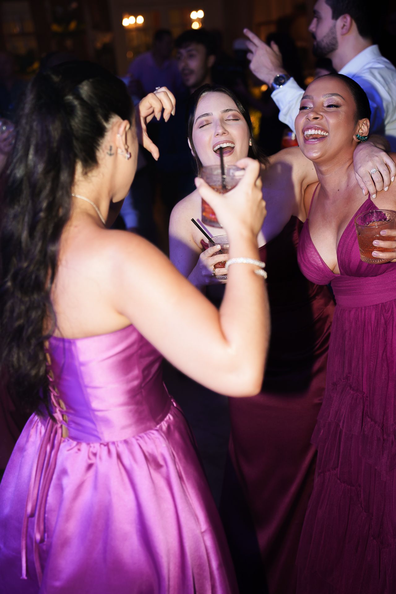 Three women in purple and burgundy formal gowns laughing and dancing at wedding reception.